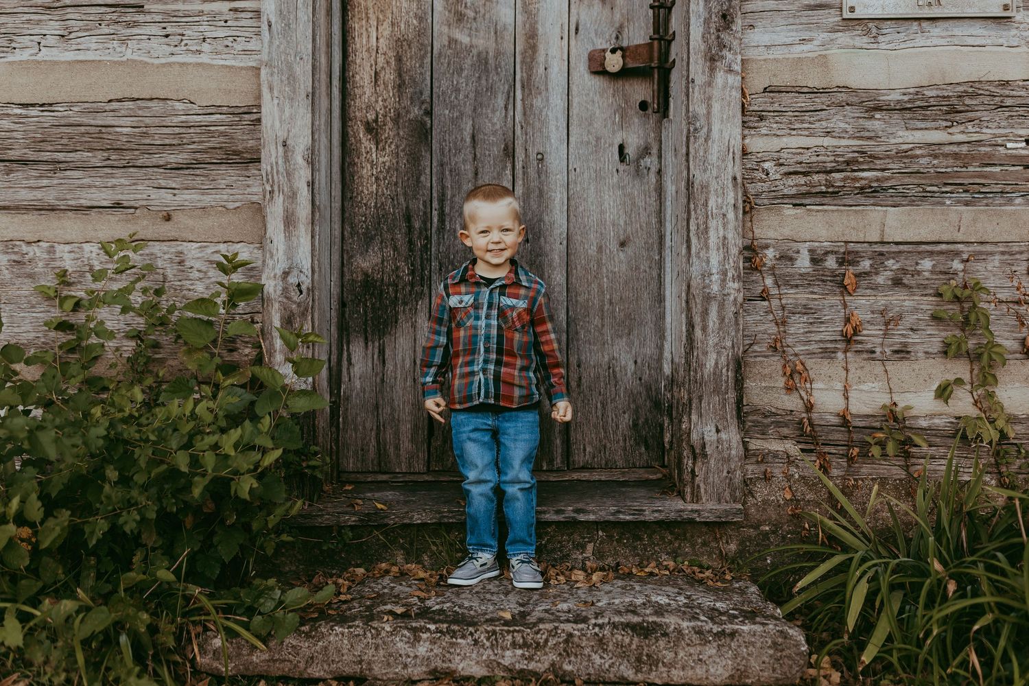 Small child standing in front of weathered wooden door wearing plaid shirt and jeans.