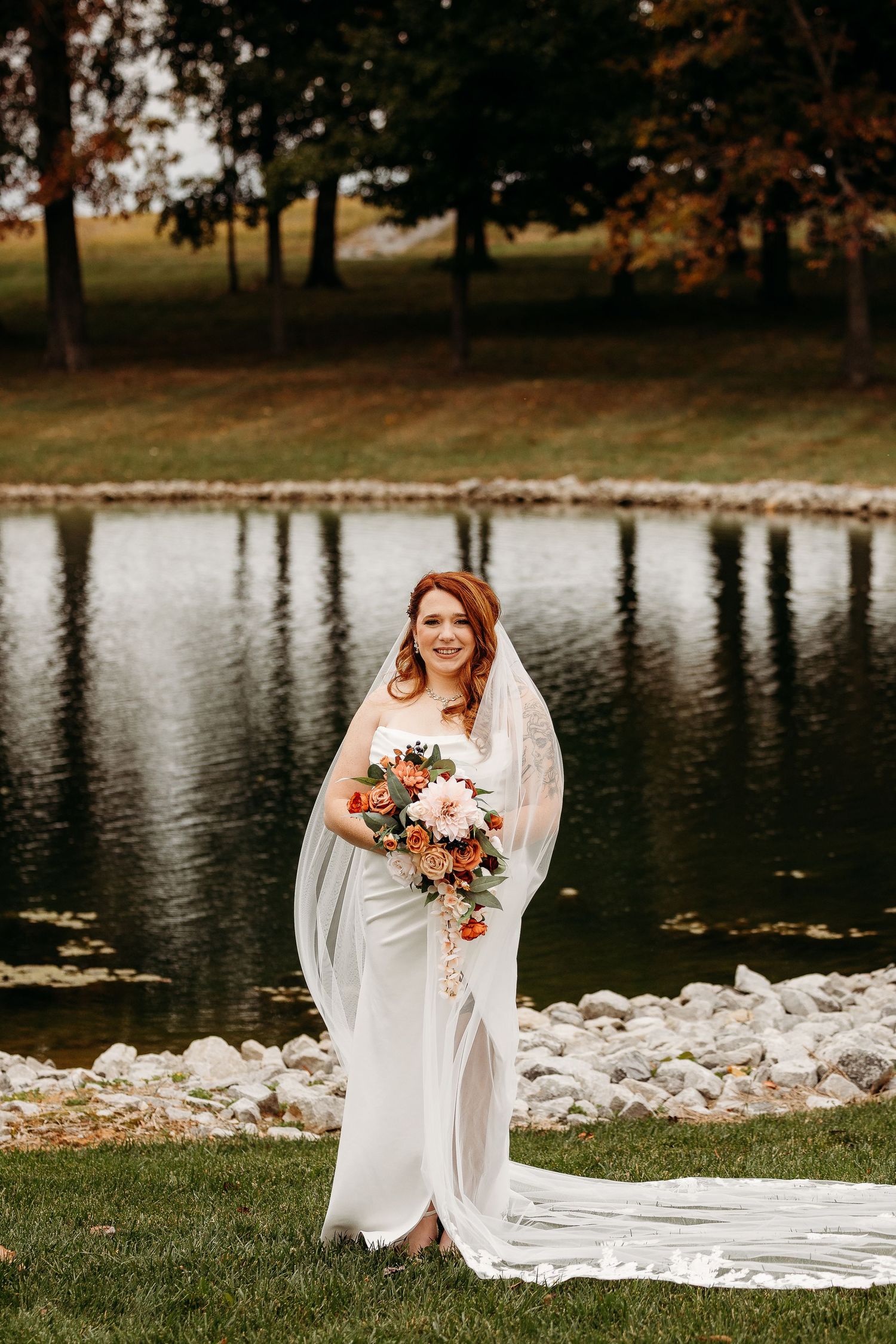 Radiant bride in white gown with veil holds orange and pink bouquet standing beside autumn pond.