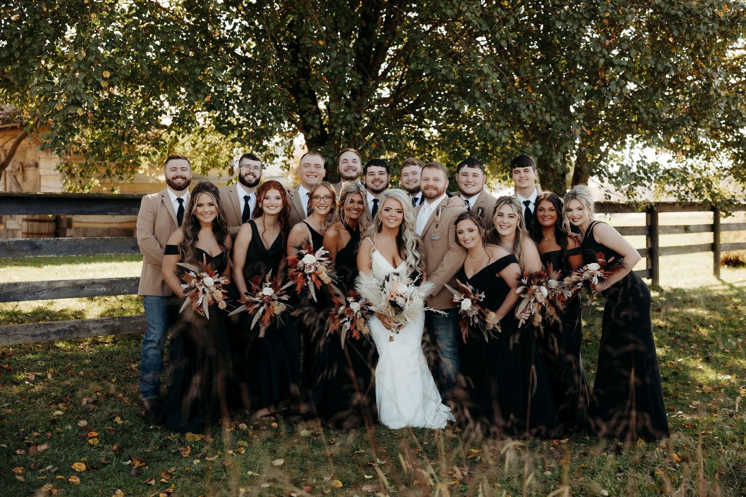 Smiling wedding party posing under autumn trees, bridesmaids holding fall bouquets in black dresses.