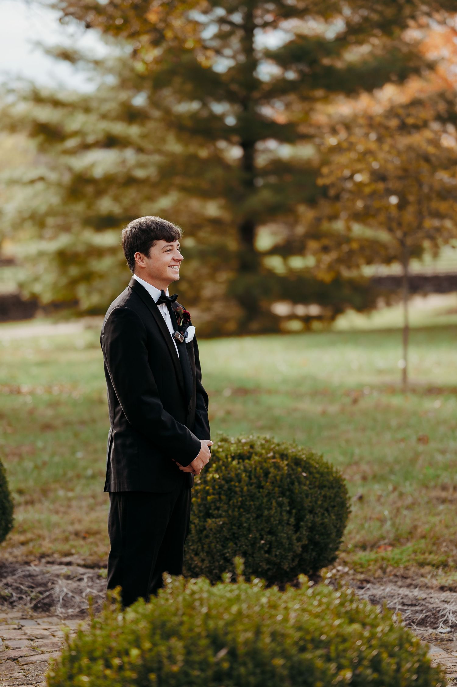 Smiling groom in black tuxedo with boutonniere standing outdoors among manicured shrubs with autumn trees in background.
