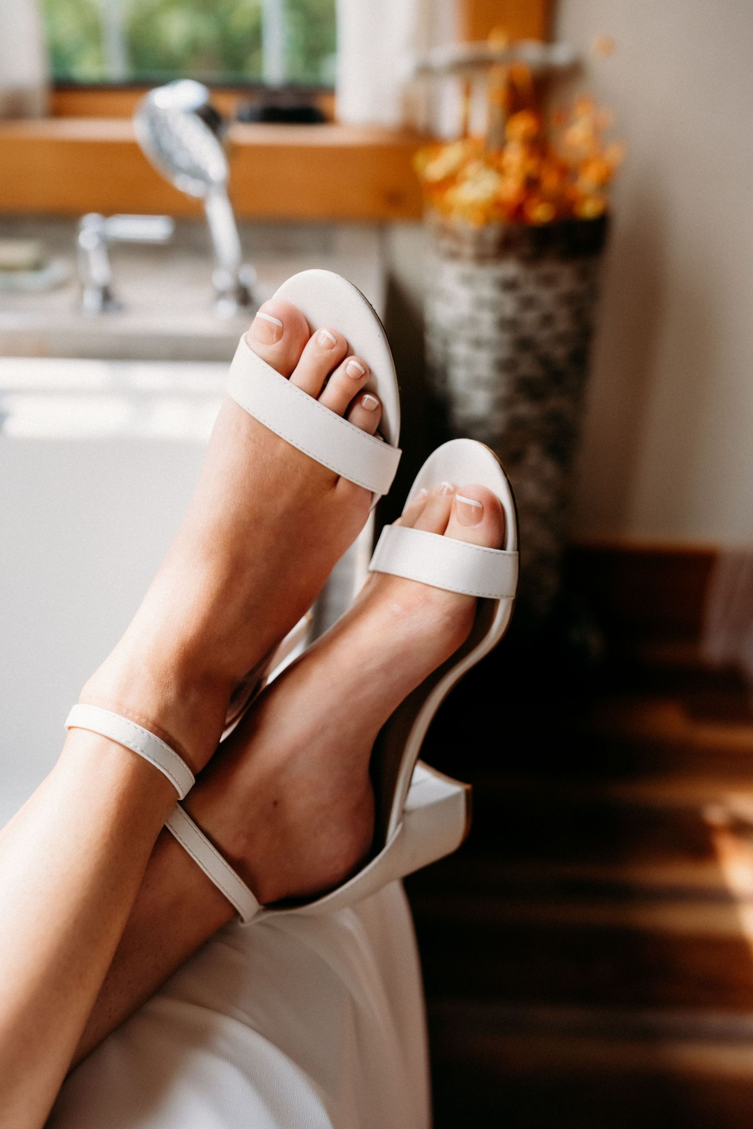 Close-up of a bride wearing elegant white block heel sandals with ankle straps, legs raised against a bathroom backdrop.
