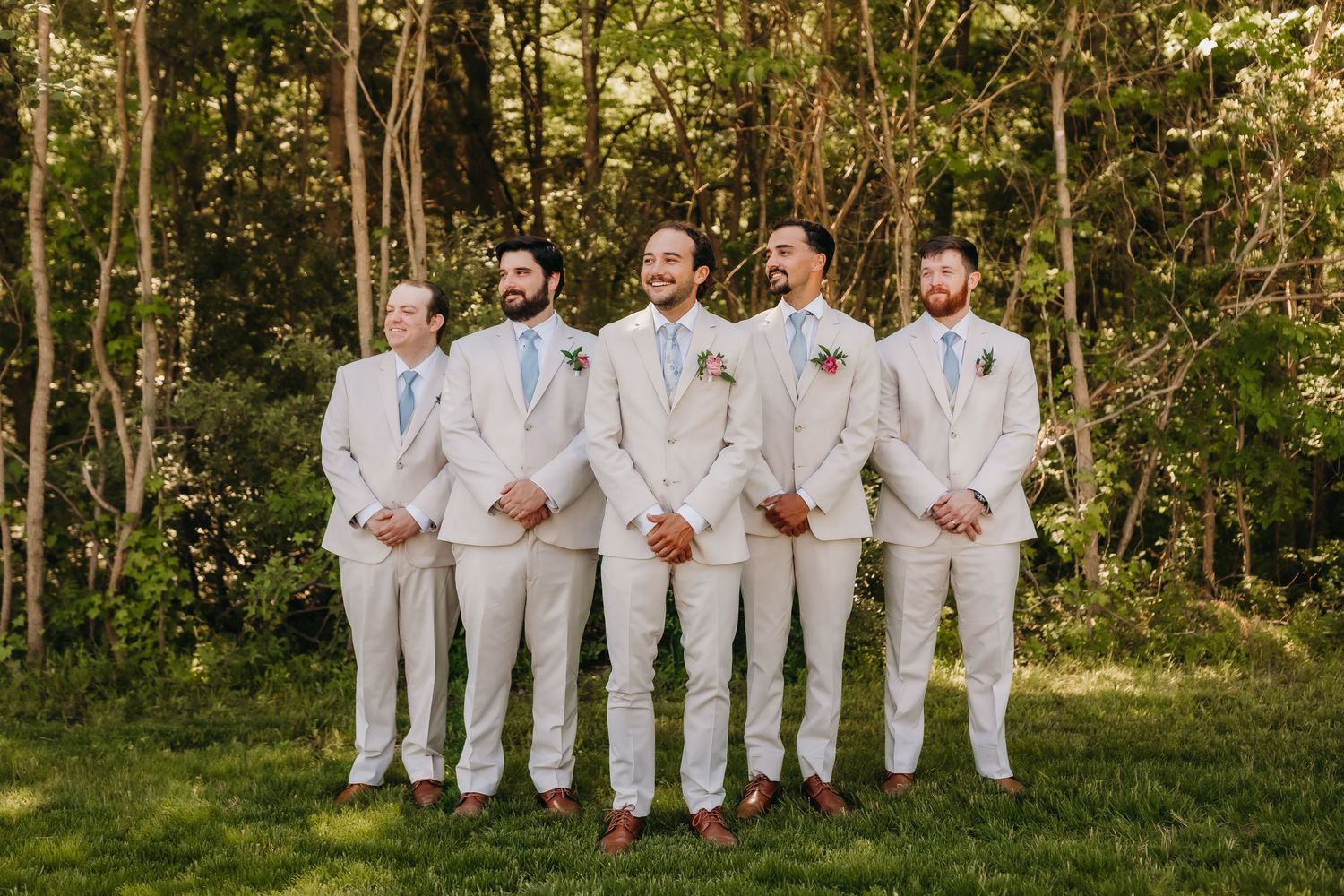 Groom and groomsmen in matching tan suits with boutonnieres posing outdoors in front of green trees.