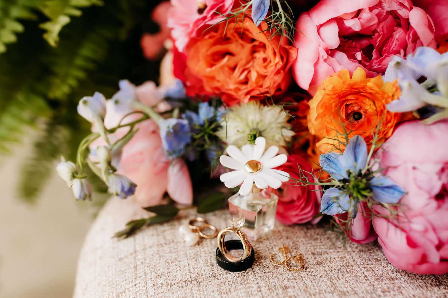 Colorful wedding bouquet with pink, orange, and blue flowers beside gold and black wedding rings on burlap.