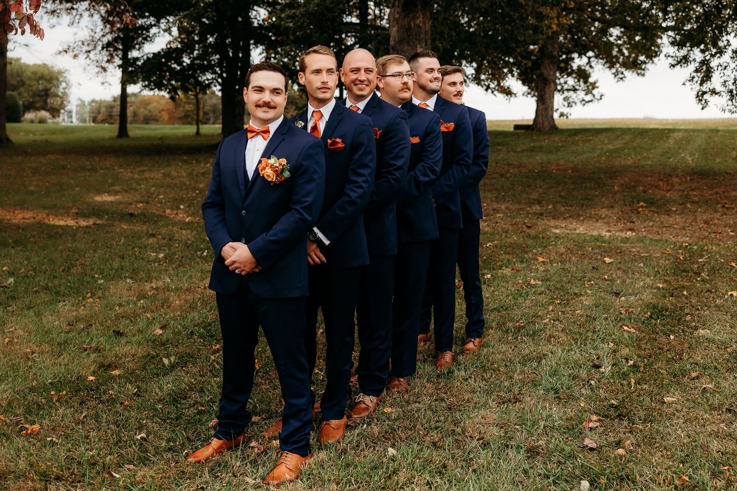 Groomsmen in navy blue suits with orange accessories line up outdoors on green grass at wedding.