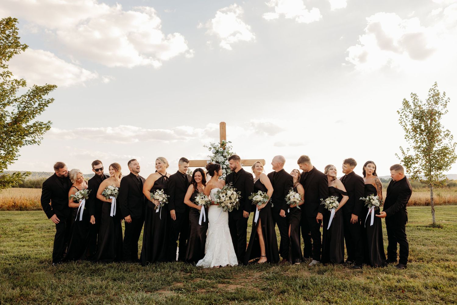 Large wedding party in black attire posing outdoors near a wooden cross on a sunny day.