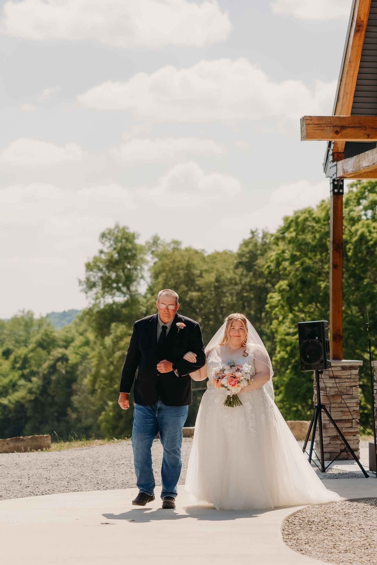 Bride in white ball gown walks with escort toward wedding venue surrounded by green trees.