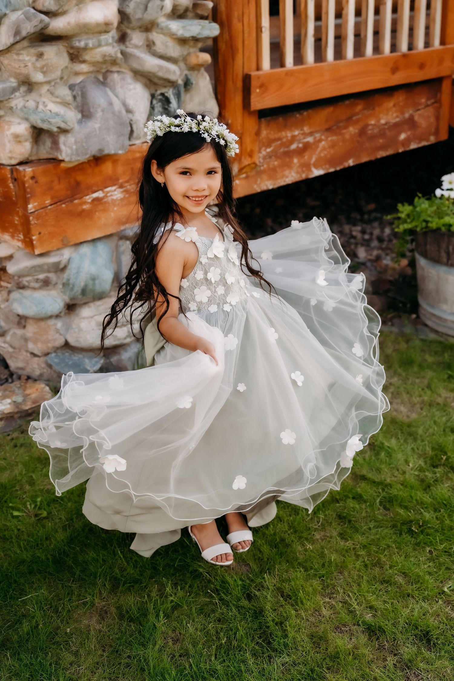 A young flower girl twirls in a white floral tulle dress with a floral crown outdoors at a wedding.