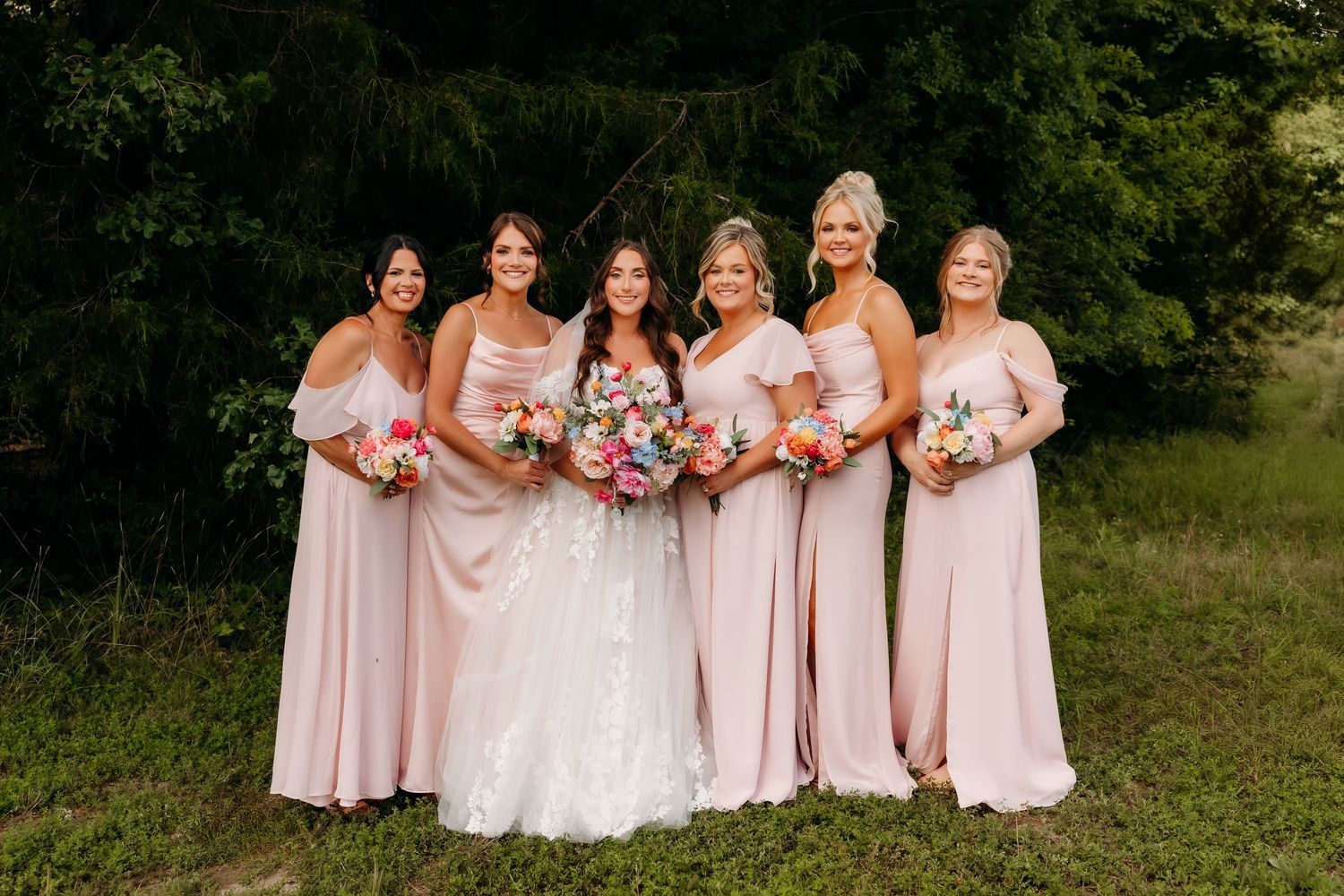 Bride in lace gown poses with five bridesmaids in blush pink dresses holding colorful bouquets.