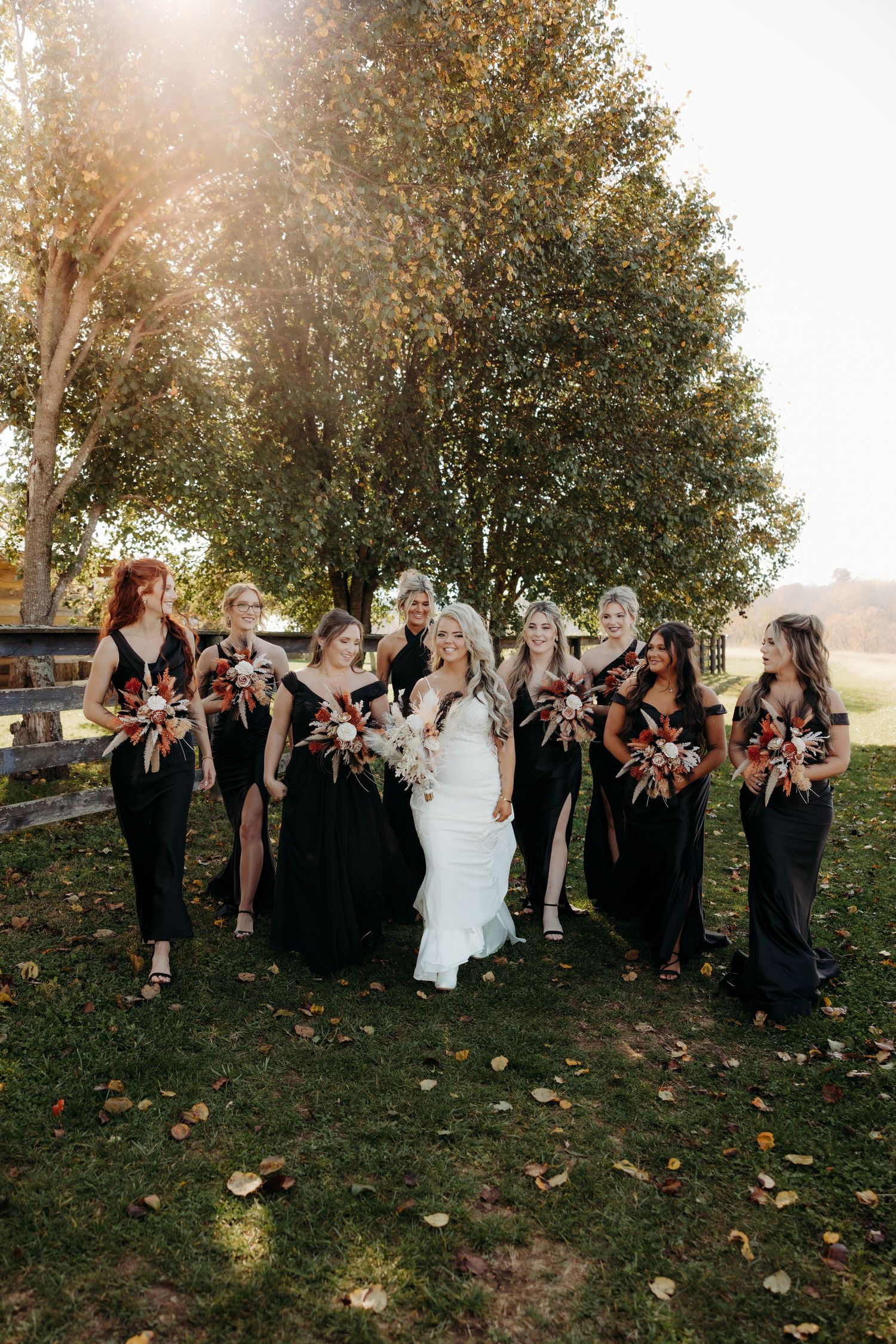 Wedding party celebrates under autumn trees with golden sunlight filtering through.