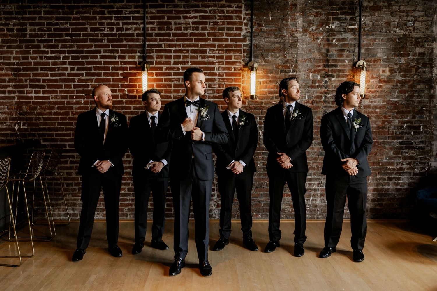 Groomsmen in black tuxedos lined up against industrial brick wall backdrop at The Frazier History Museum in Louisville, KY.