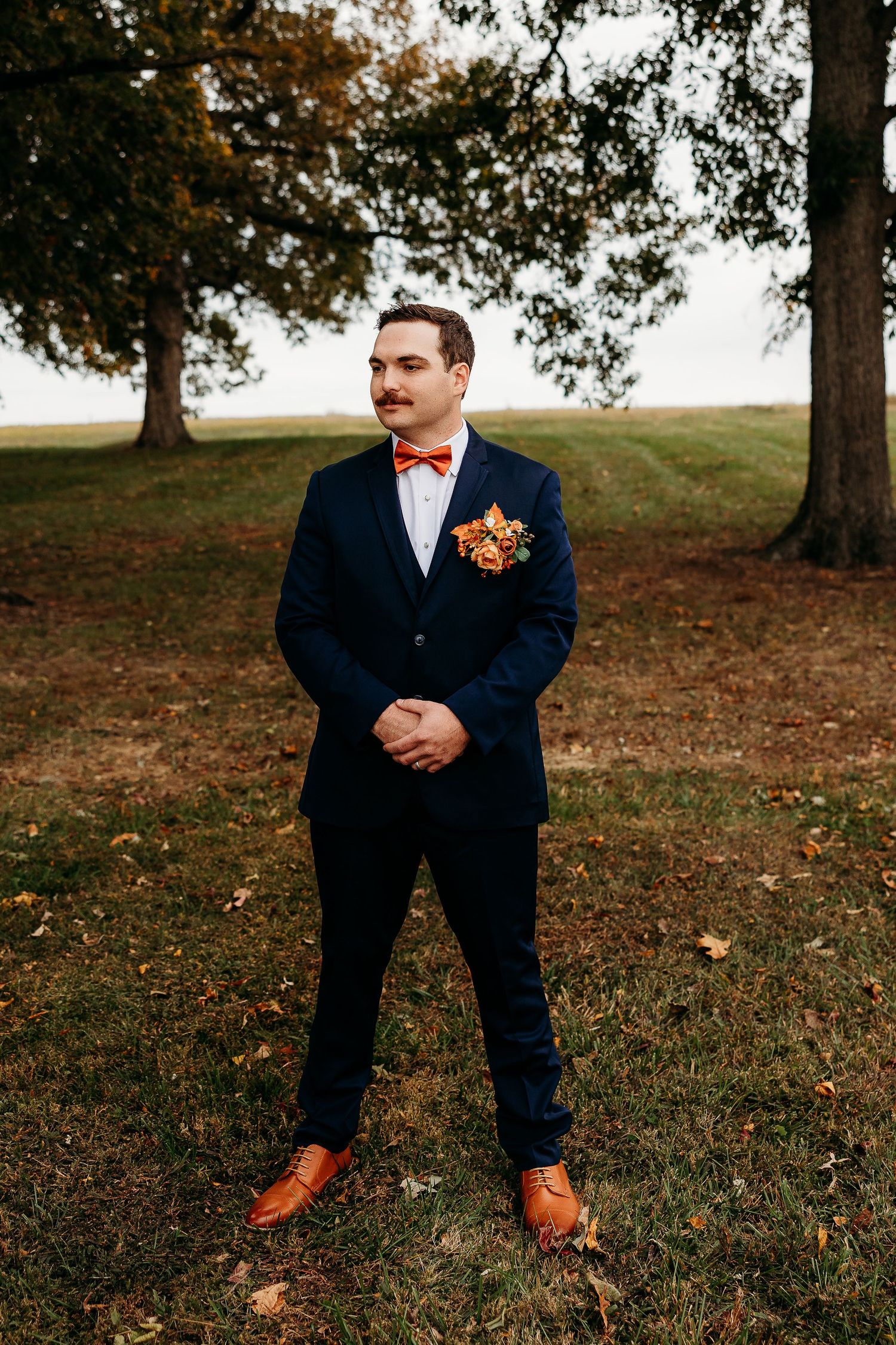 Groom in navy suit with coral boutonniere poses on autumn grass with fallen leaves.