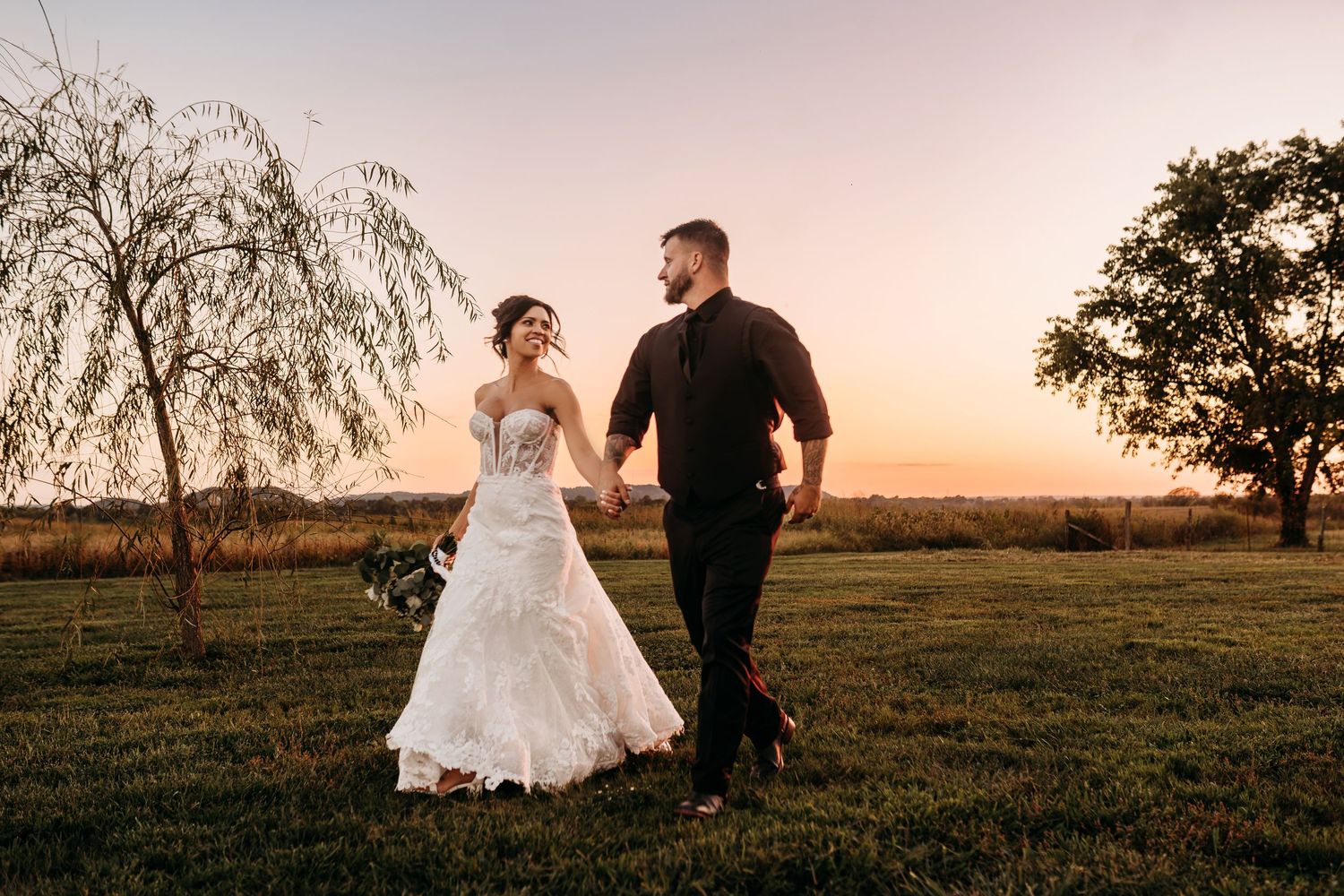 Bride and groom holding hands walking at sunset on green lawn, wedding dress and black attire, golden sky backdrop.