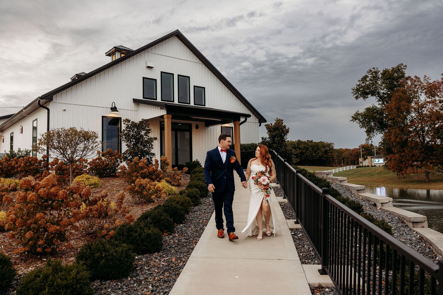 Bride and groom walk hand in hand outside a modern white barn wedding venue on a cloudy day.