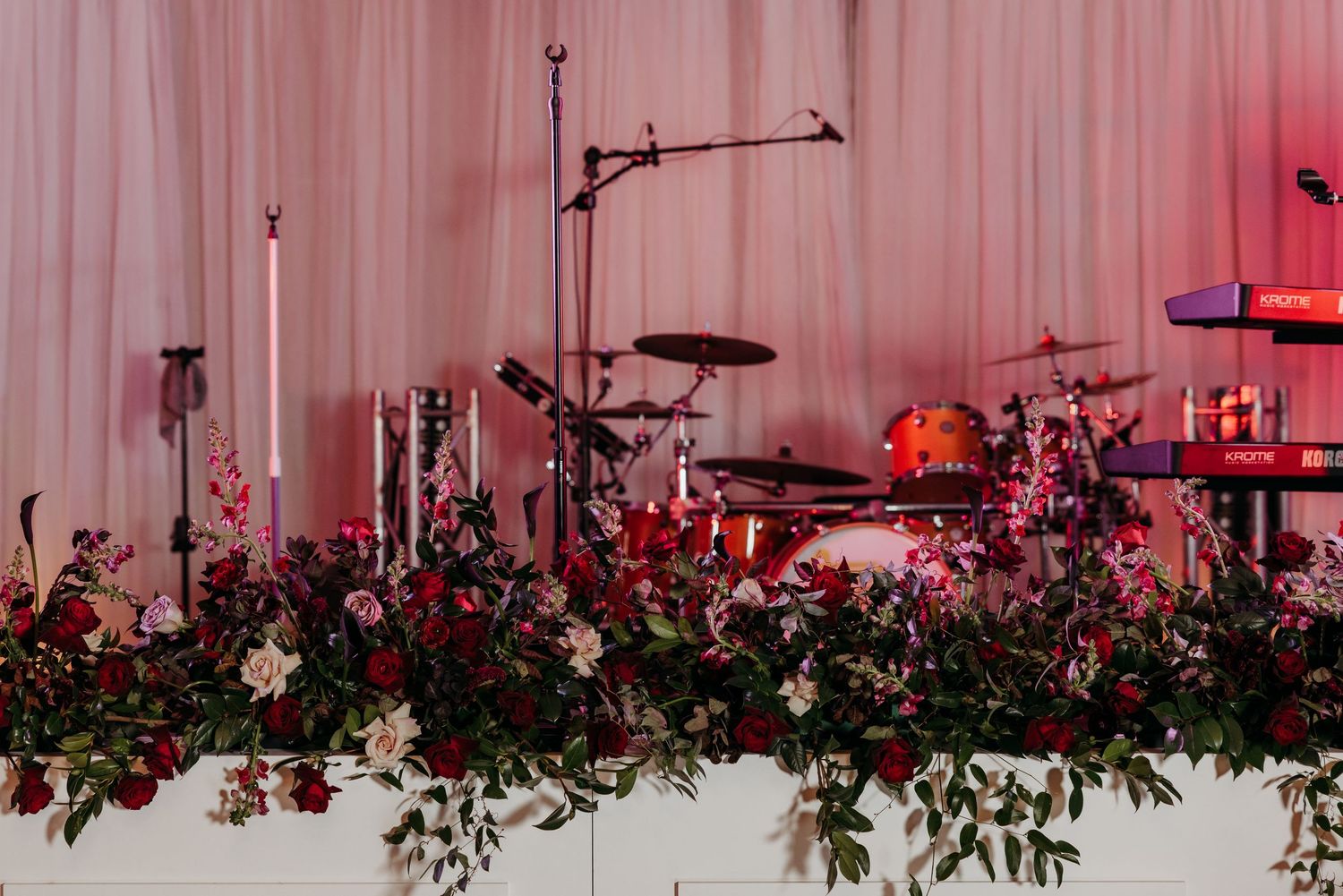 Elegant wedding stage with lush red floral arrangements, candles, and a drum set against pink draped backdrop.