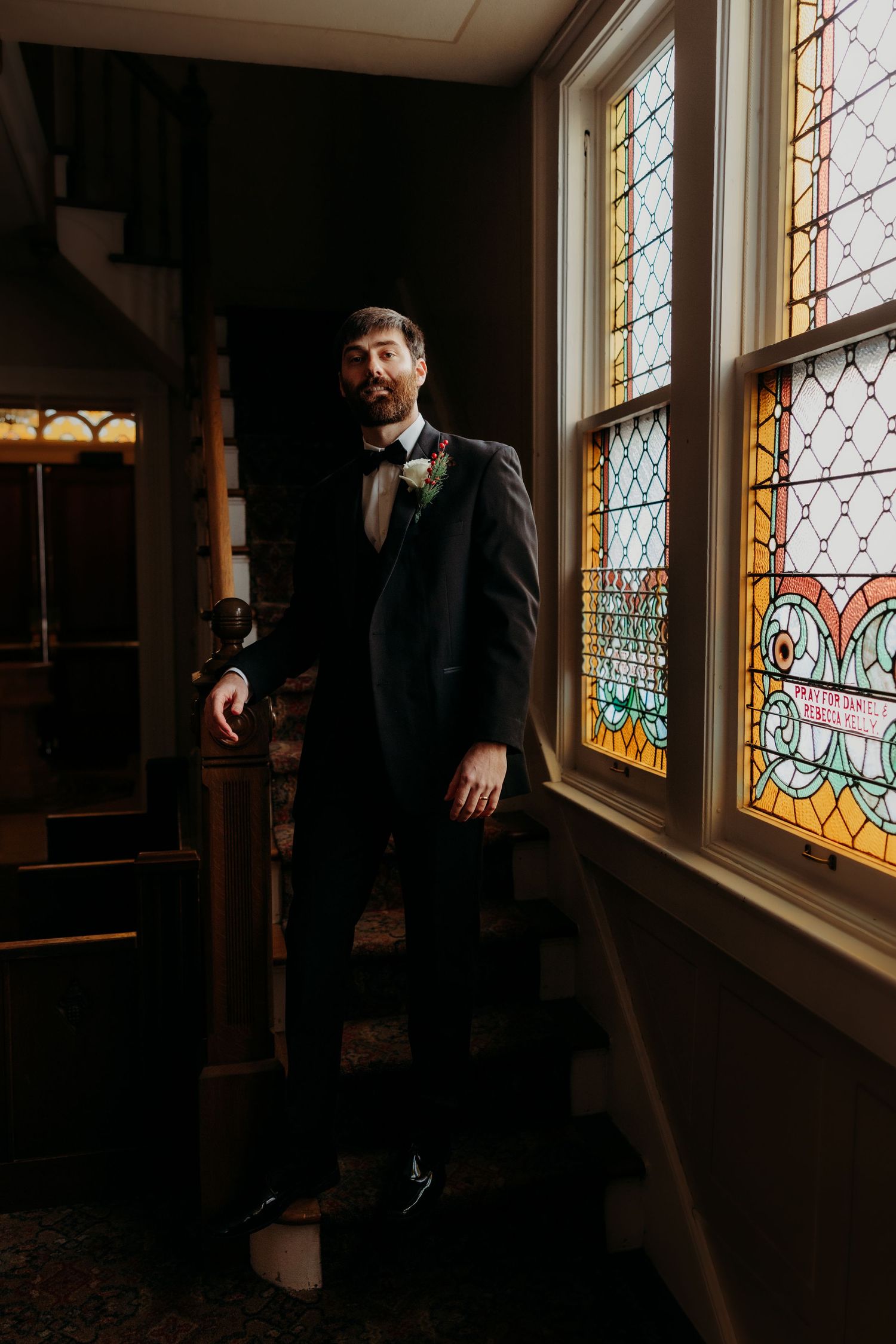 Dapper groom in black tuxedo standing by colorful stained glass church windows.