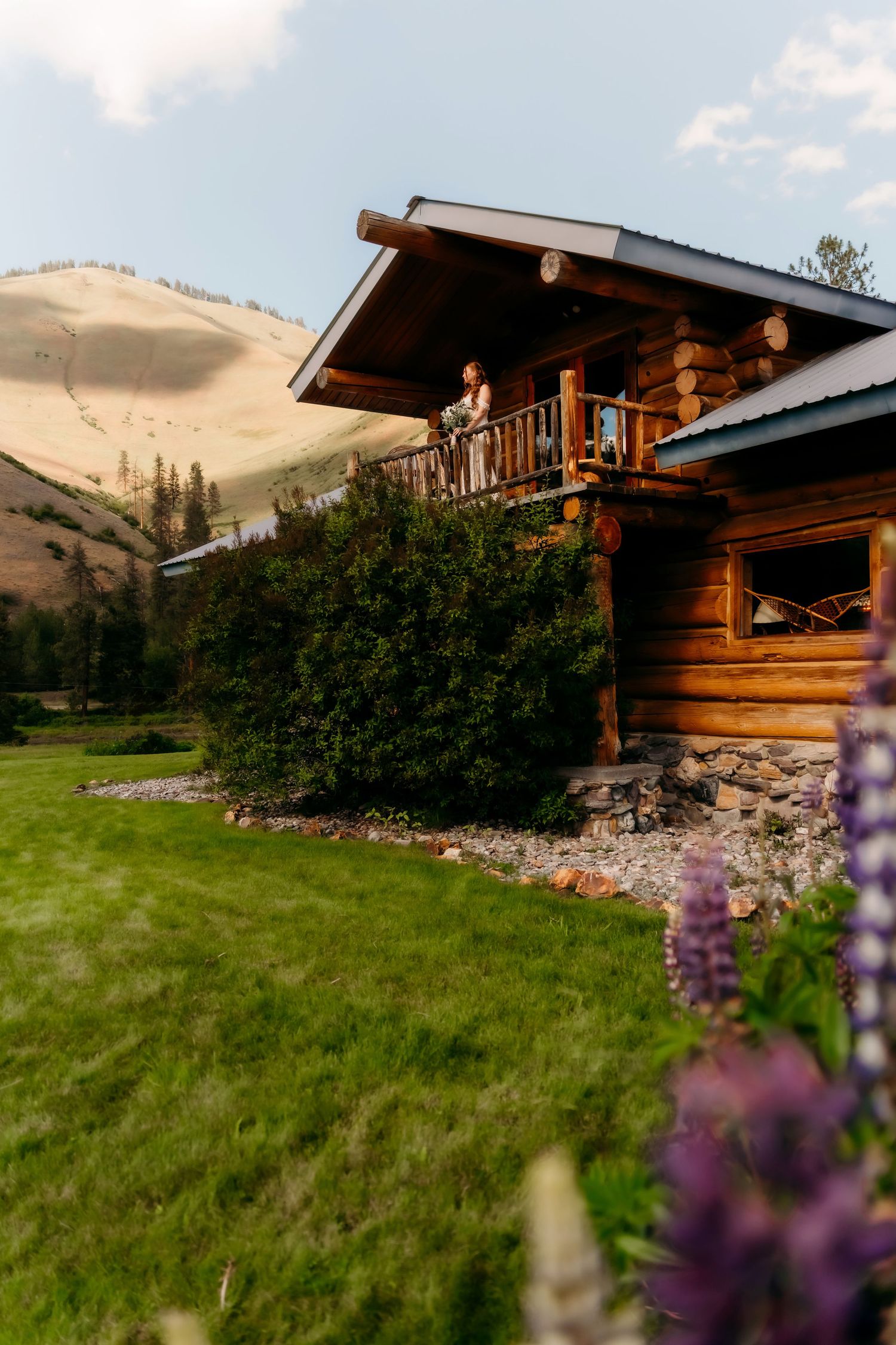 Rustic log cabin with wraparound balcony surrounded by lupine flowers and mountain landscape at dusk.