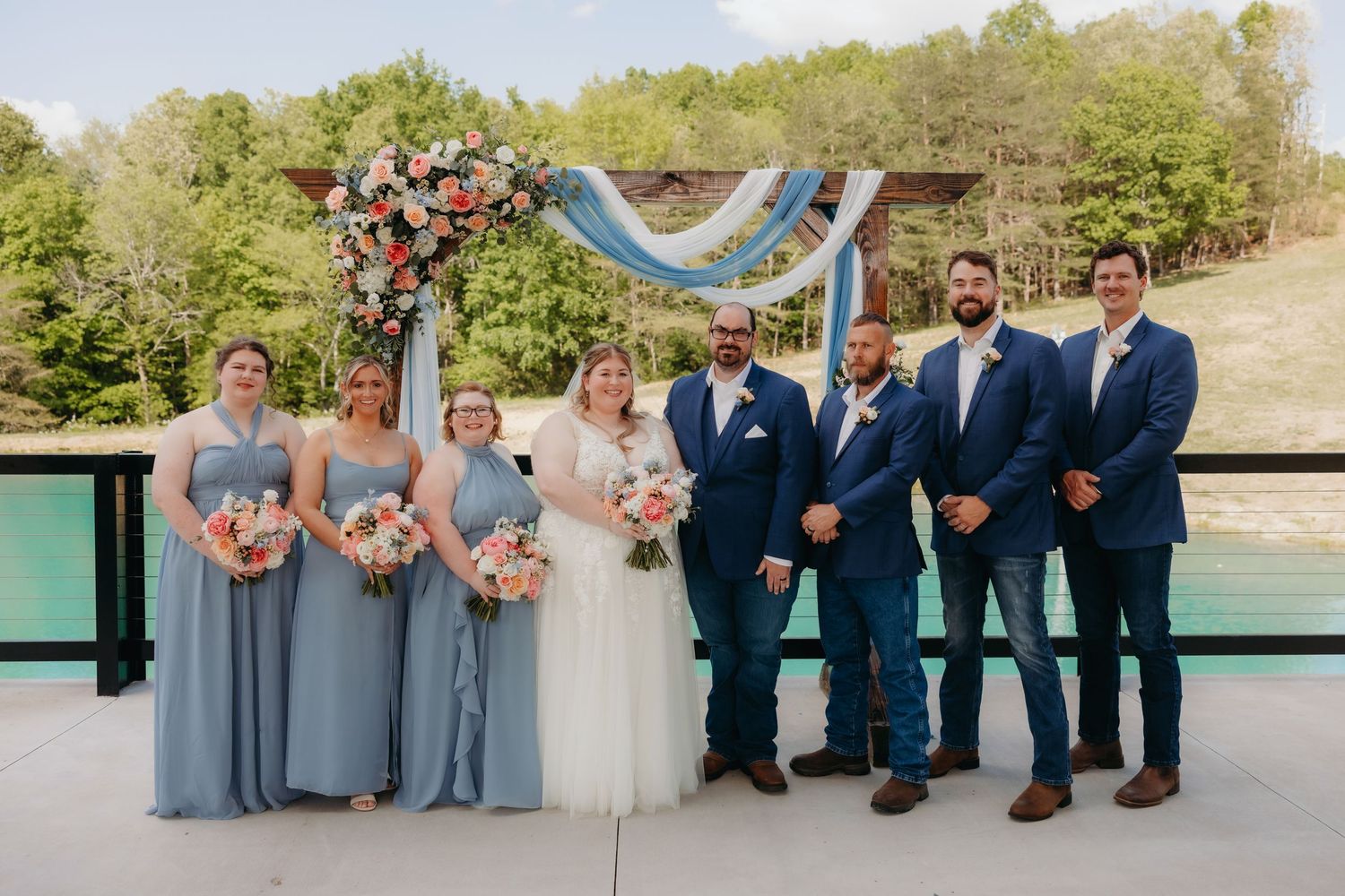 Wedding party poses outdoors on a deck with bridesmaids in blue gowns and groomsmen in navy suits under a floral arch.