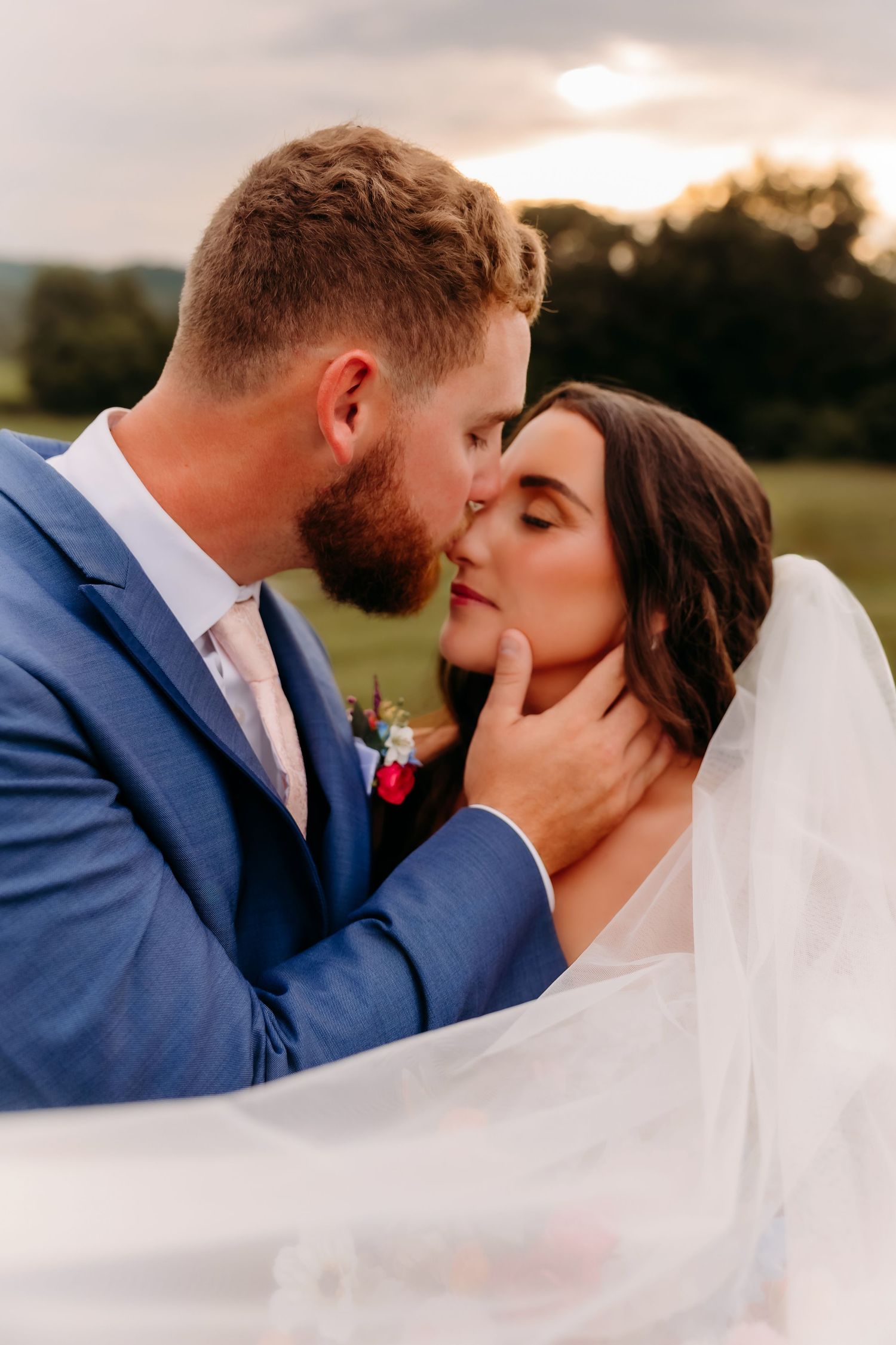 Groom in blue suit kisses bride's forehead while cradling her face, veil flowing at golden hour outdoors.