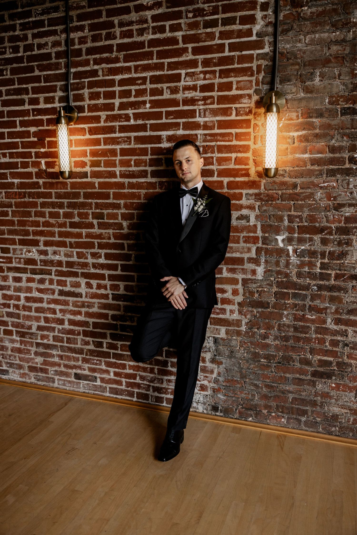 Groom poses formally against exposed brick wall in classic black tuxedo.