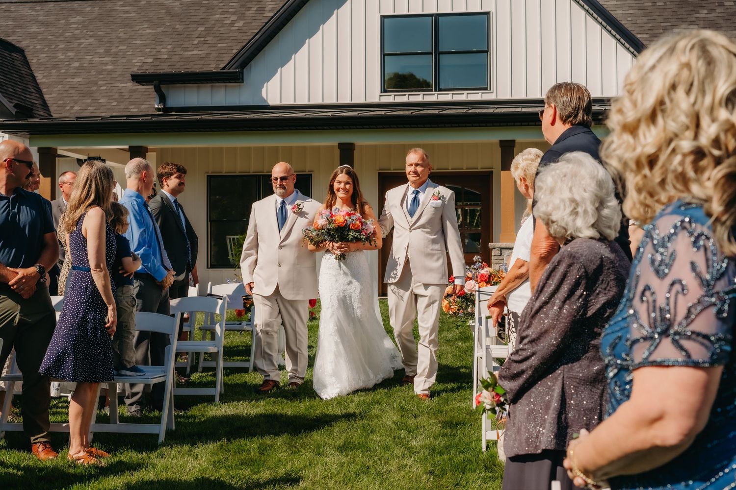 Bride walking down outdoor aisle with escort past seated guests at sunny barn wedding ceremony.