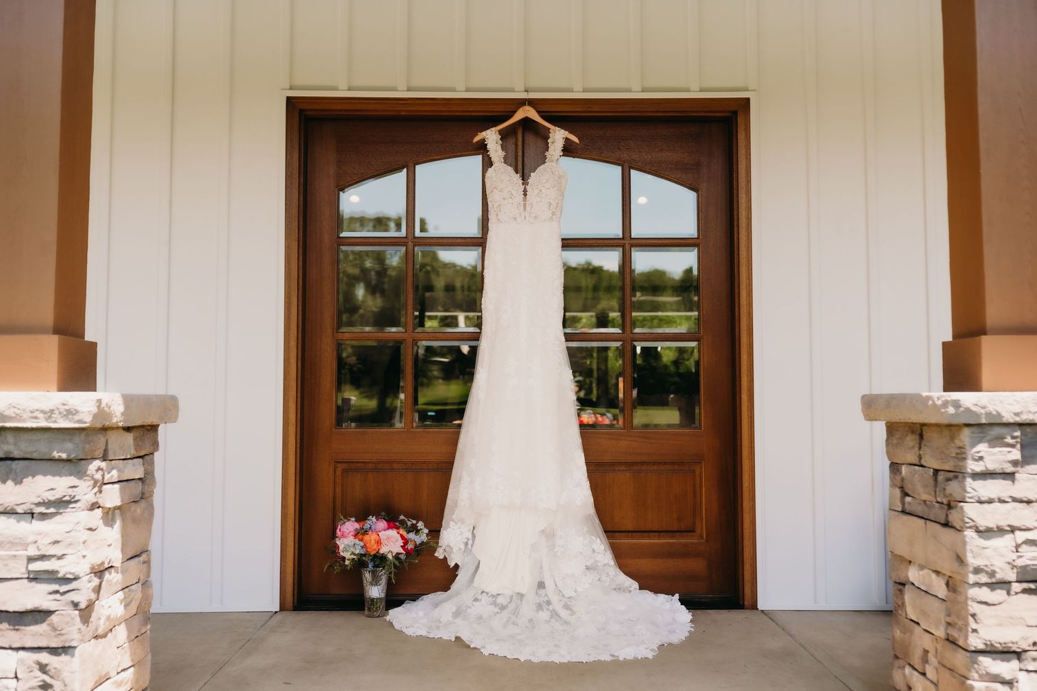 Elegant lace wedding dress hanging on wooden double doors of a rustic venue with stone pillars.