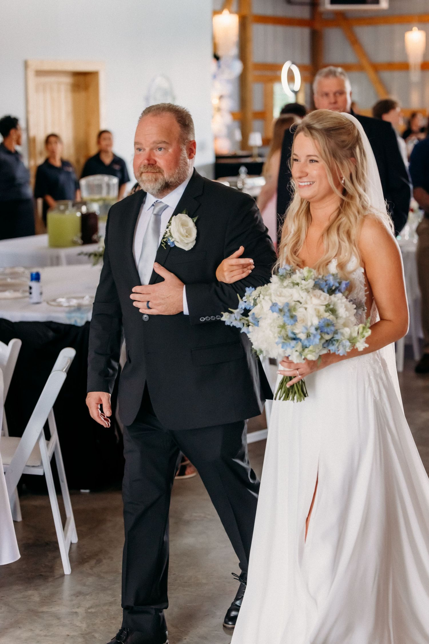Father of the bride walking his smiling daughter down the aisle in a rustic wedding venue.