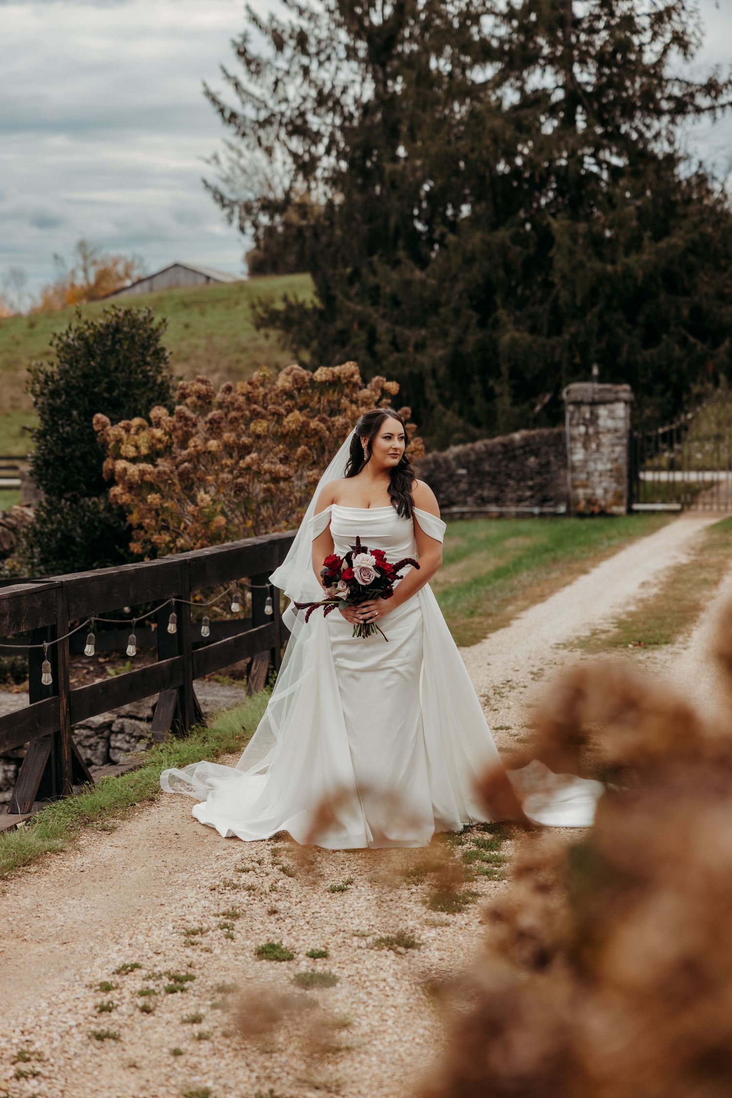 Bride in off-shoulder white gown holding red bouquet on rustic gravel path surrounded by autumn foliage.