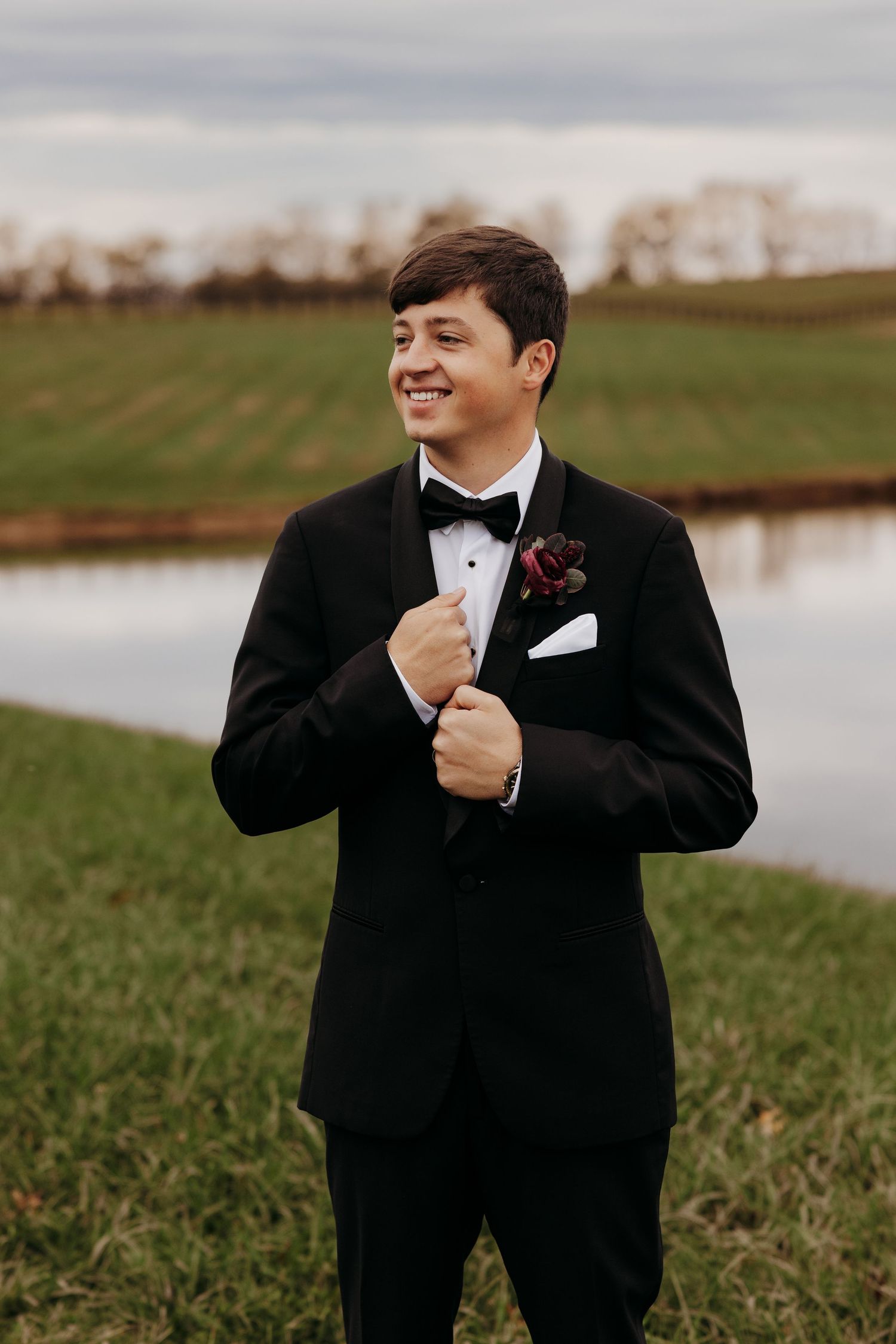 Groom in classic black tuxedo smiling outdoors by a lake during autumn.