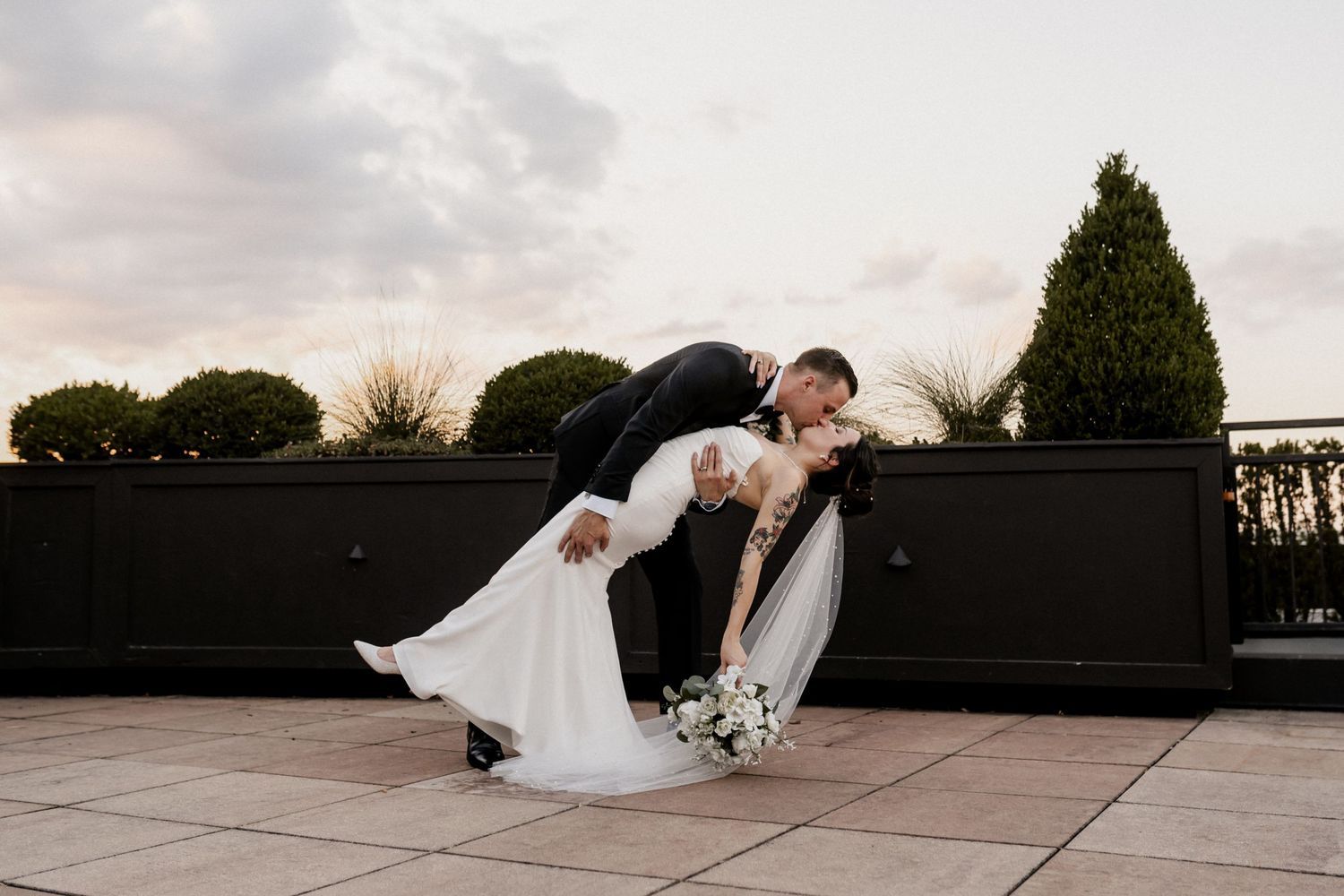 A romantic dip pose between a couple in wedding attire on an outdoor terrace at sunset.