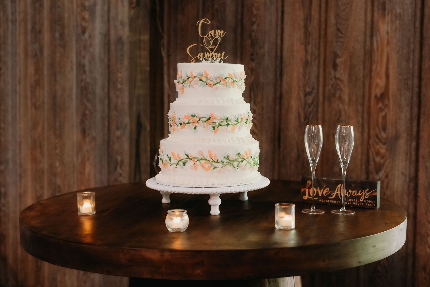 Three-tier white wedding cake with floral details and gold topper on a round wooden table.