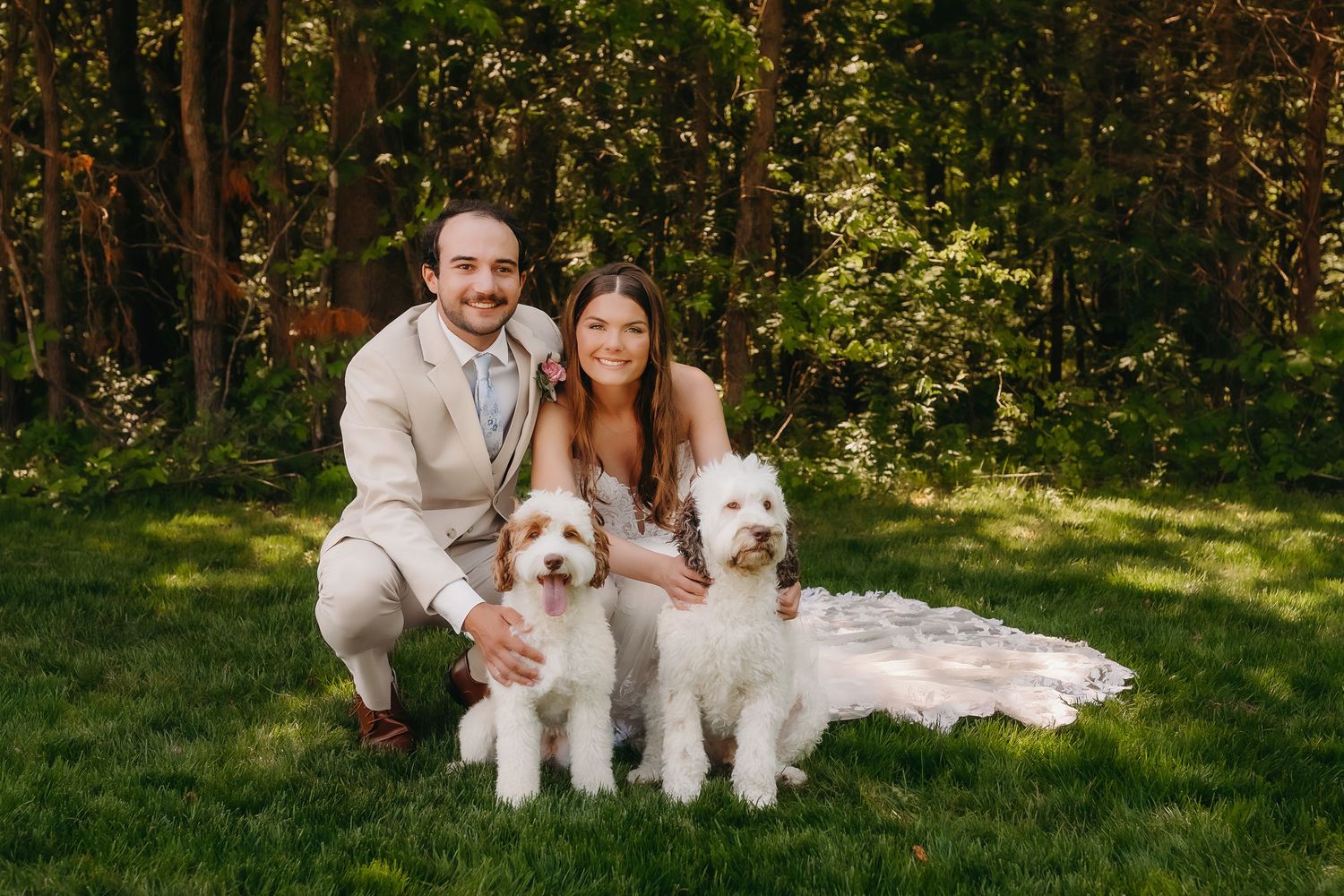Bride and groom kneeling on grass with two fluffy white dogs in front of lush green trees on wedding day.