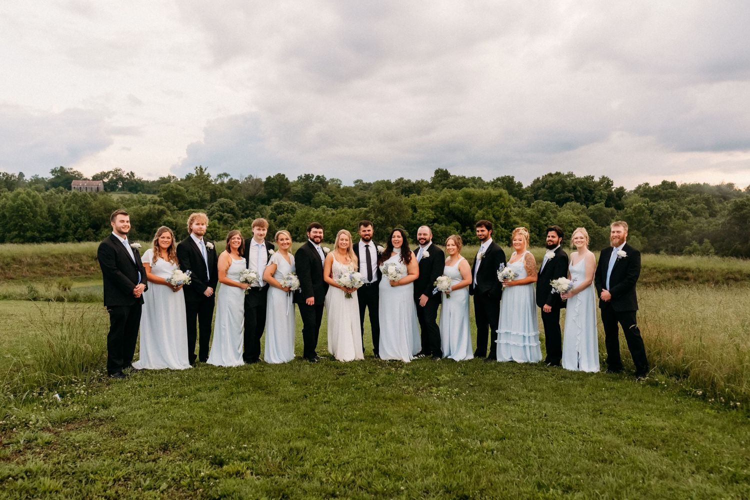 Large wedding party posing together in white dresses and black tuxedos on a green field with trees in background.