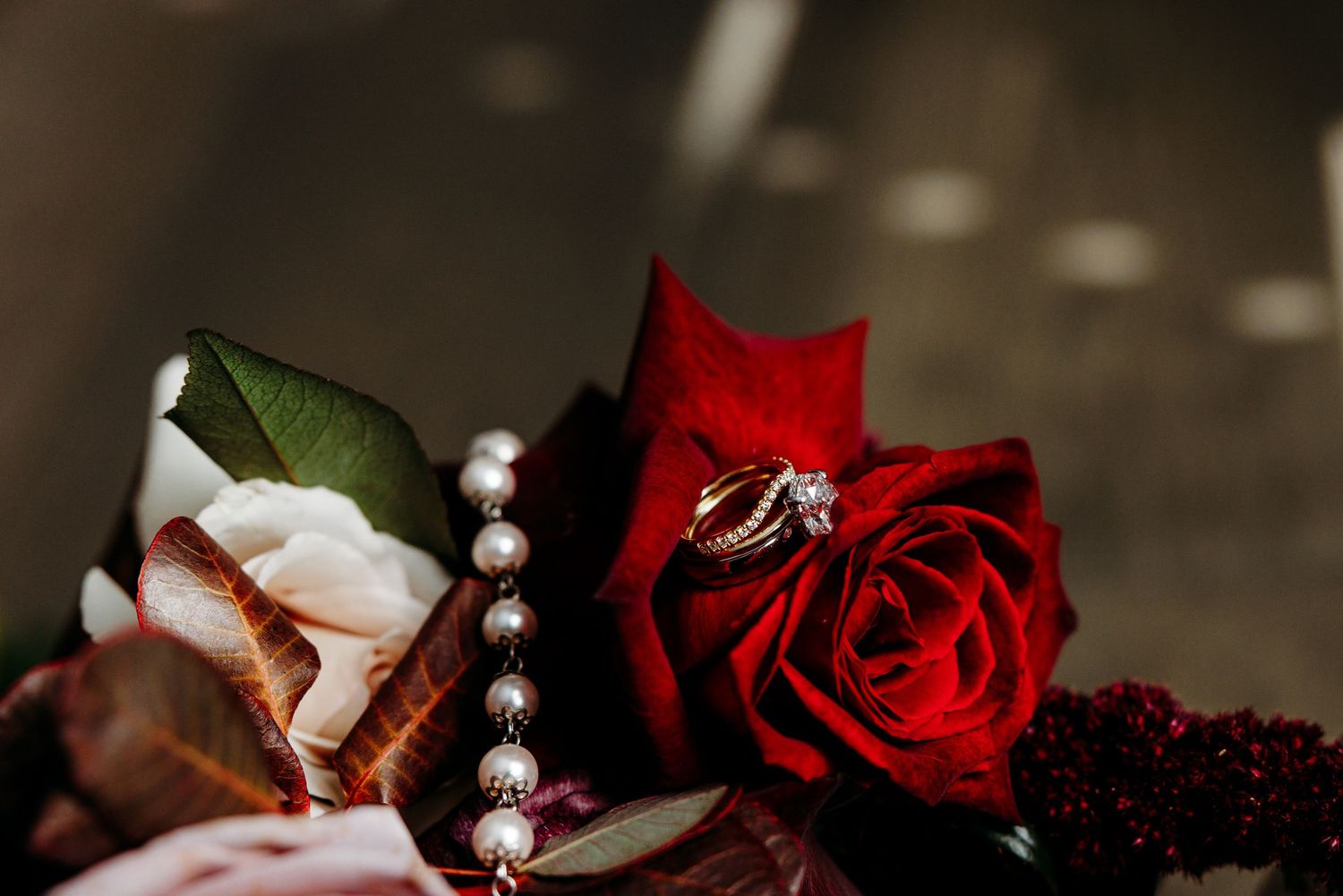 Gold engagement ring resting on a red rose surrounded by pearl necklace and floral wedding accessories.
