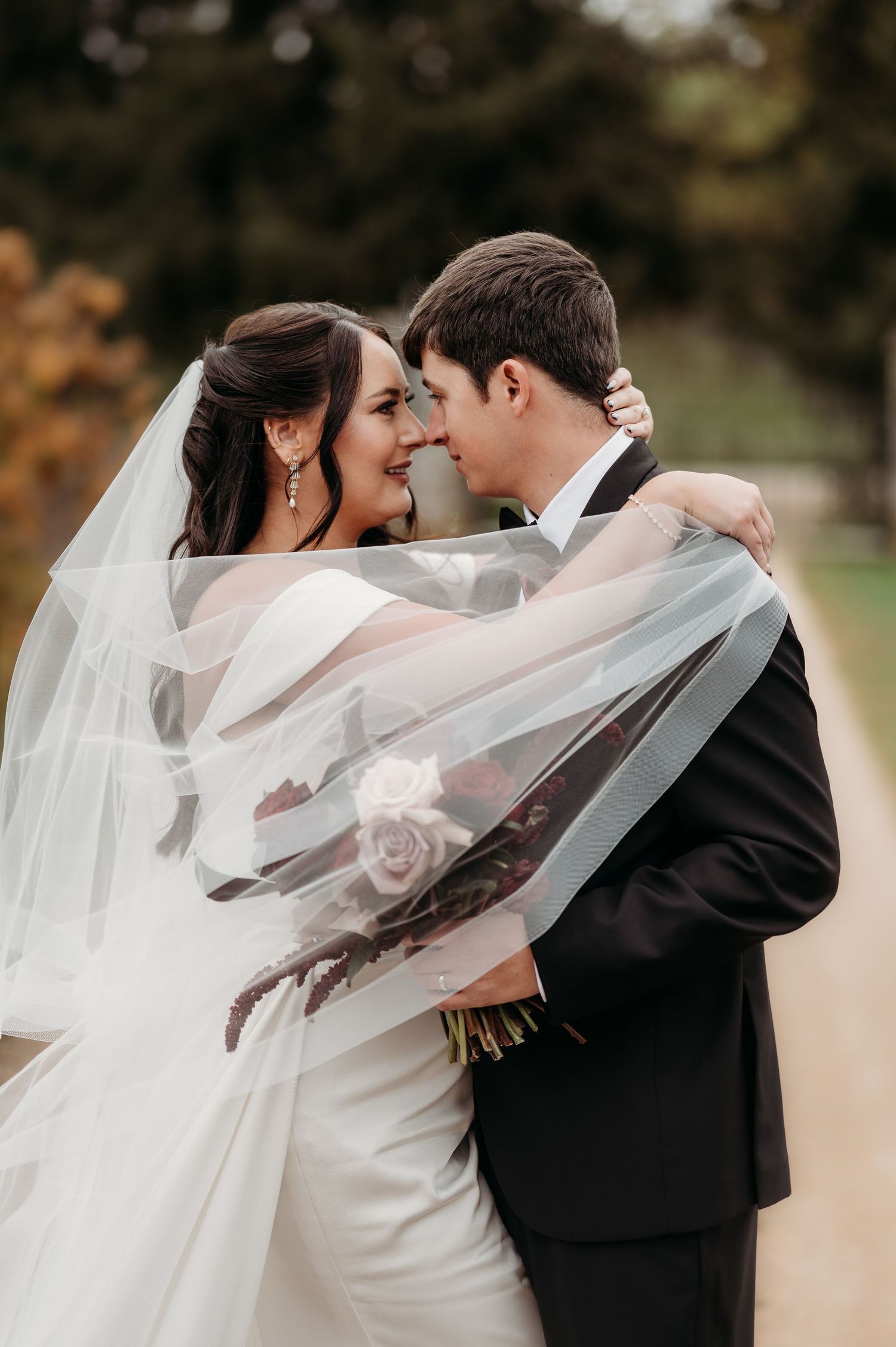 Bride and groom share a romantic moment under flowing veil, bride holding bouquet outdoors on wedding day.