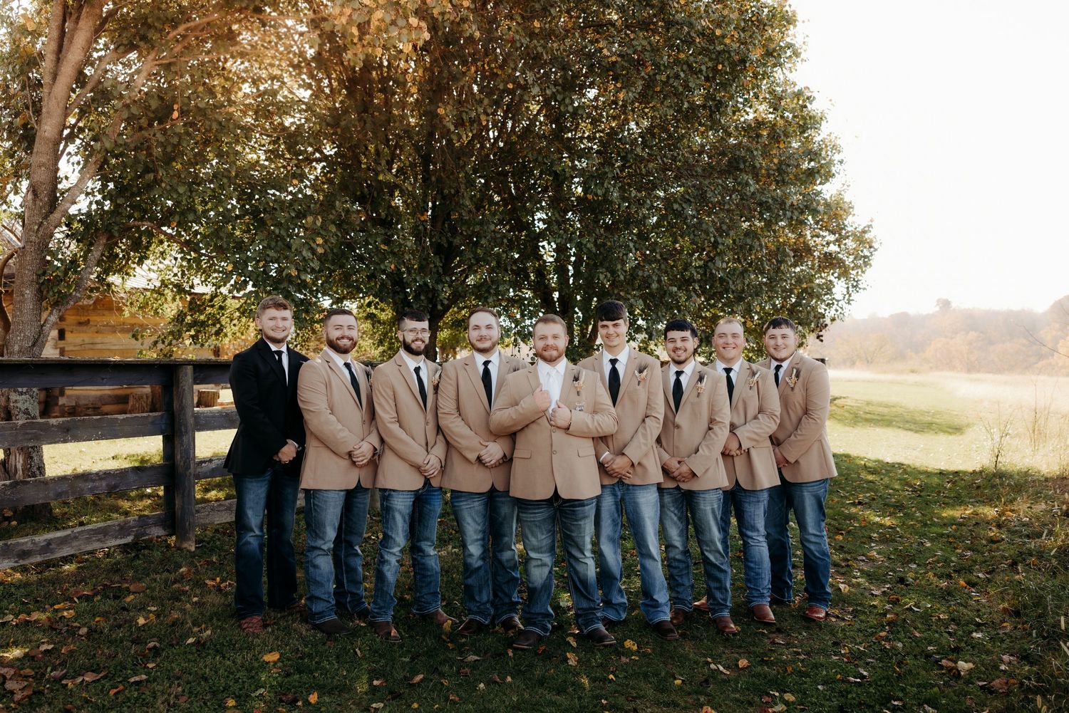 Groomsmen in matching tan blazers, white shirts, and jeans posing outdoors near a wooden fence.