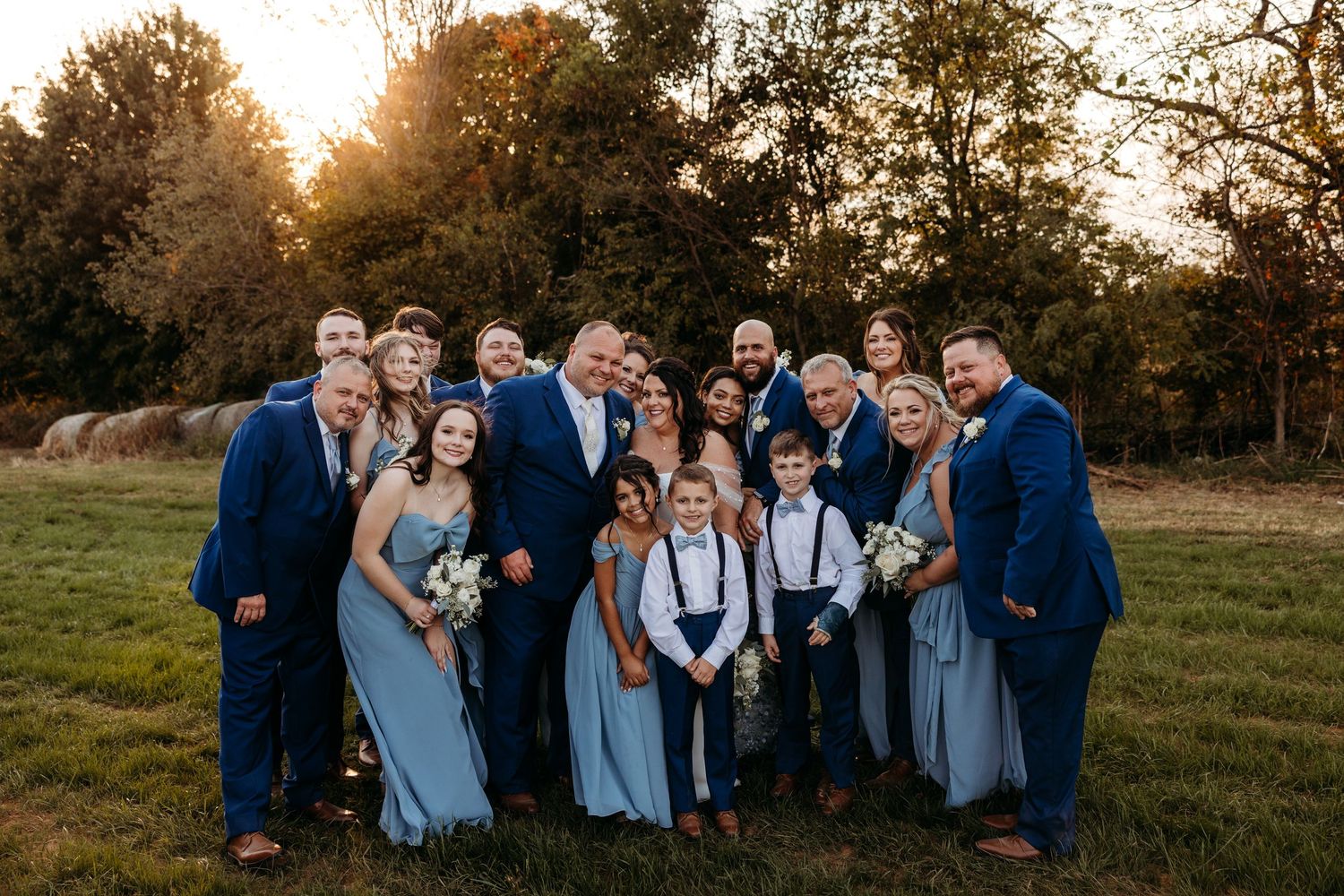 Wedding party in blue attire poses outdoors at golden hour with trees and hay bales in background.