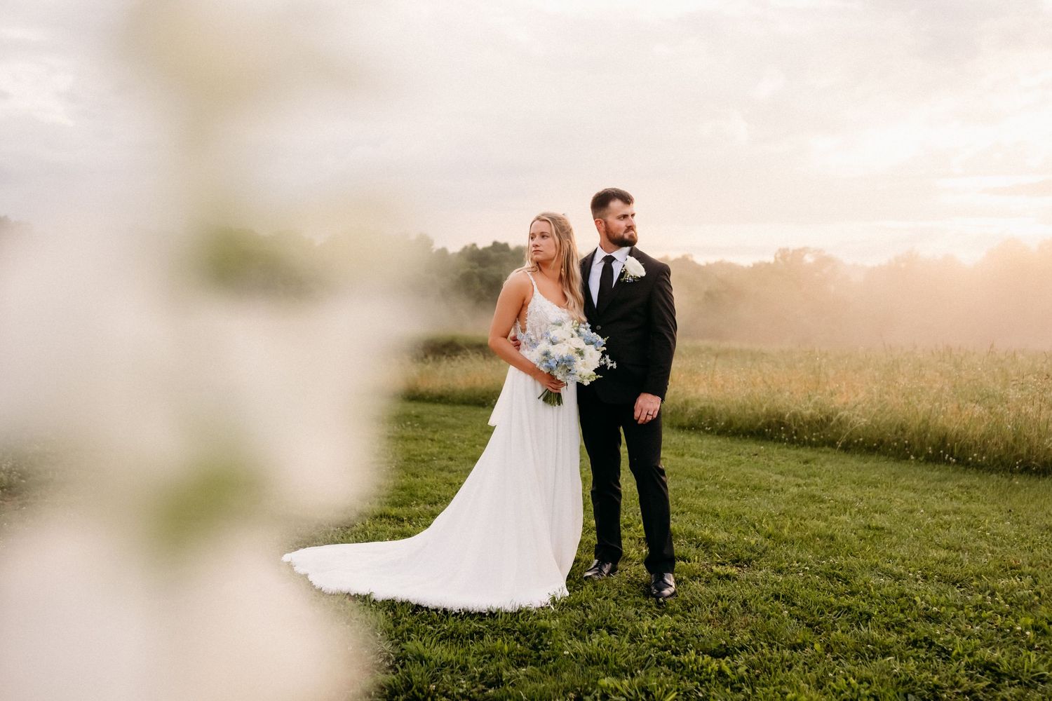 Bride and groom stand in a sunlit green field at golden hour, bride in white gown holding bouquet, groom in black tux.