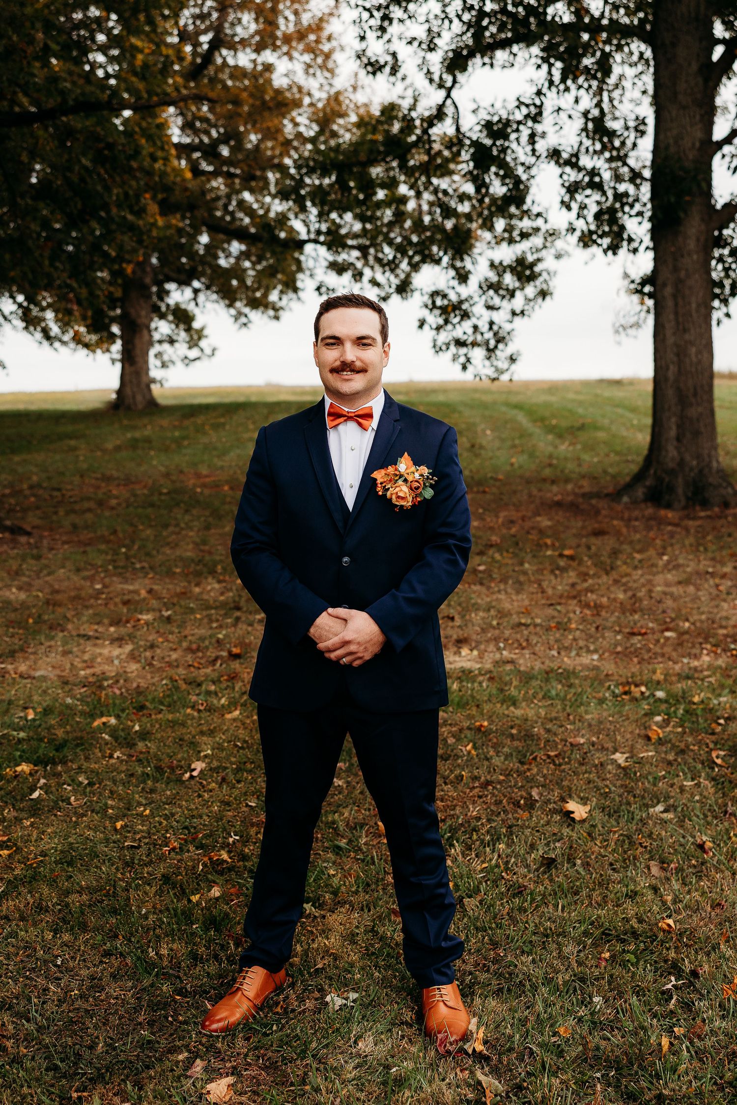 Dapper groom in navy suit with orange bow tie and boutonniere stands confidently outdoors among autumn trees.