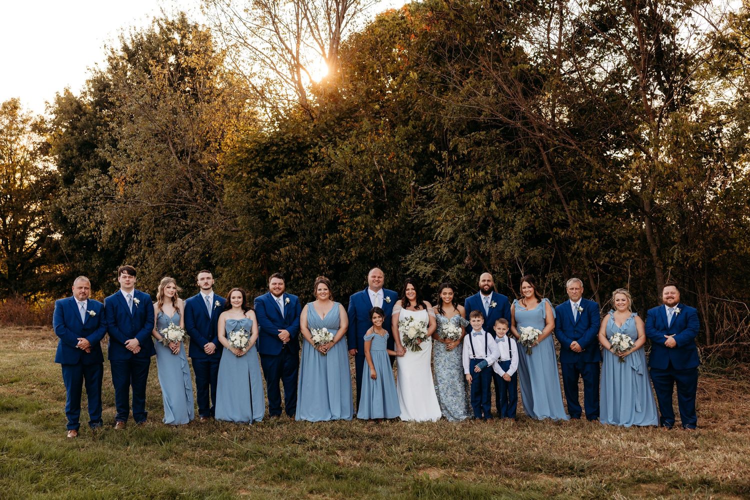 Wedding party in blue attire posing outdoors at golden hour with lush green trees in the background.