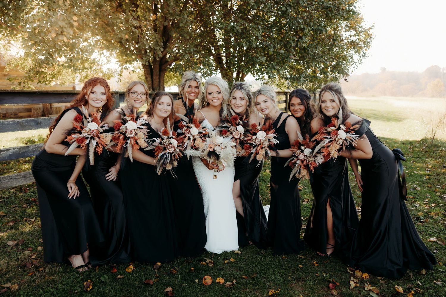 Bride in white gown with bridesmaids in black dresses holding fall bouquets outdoors.