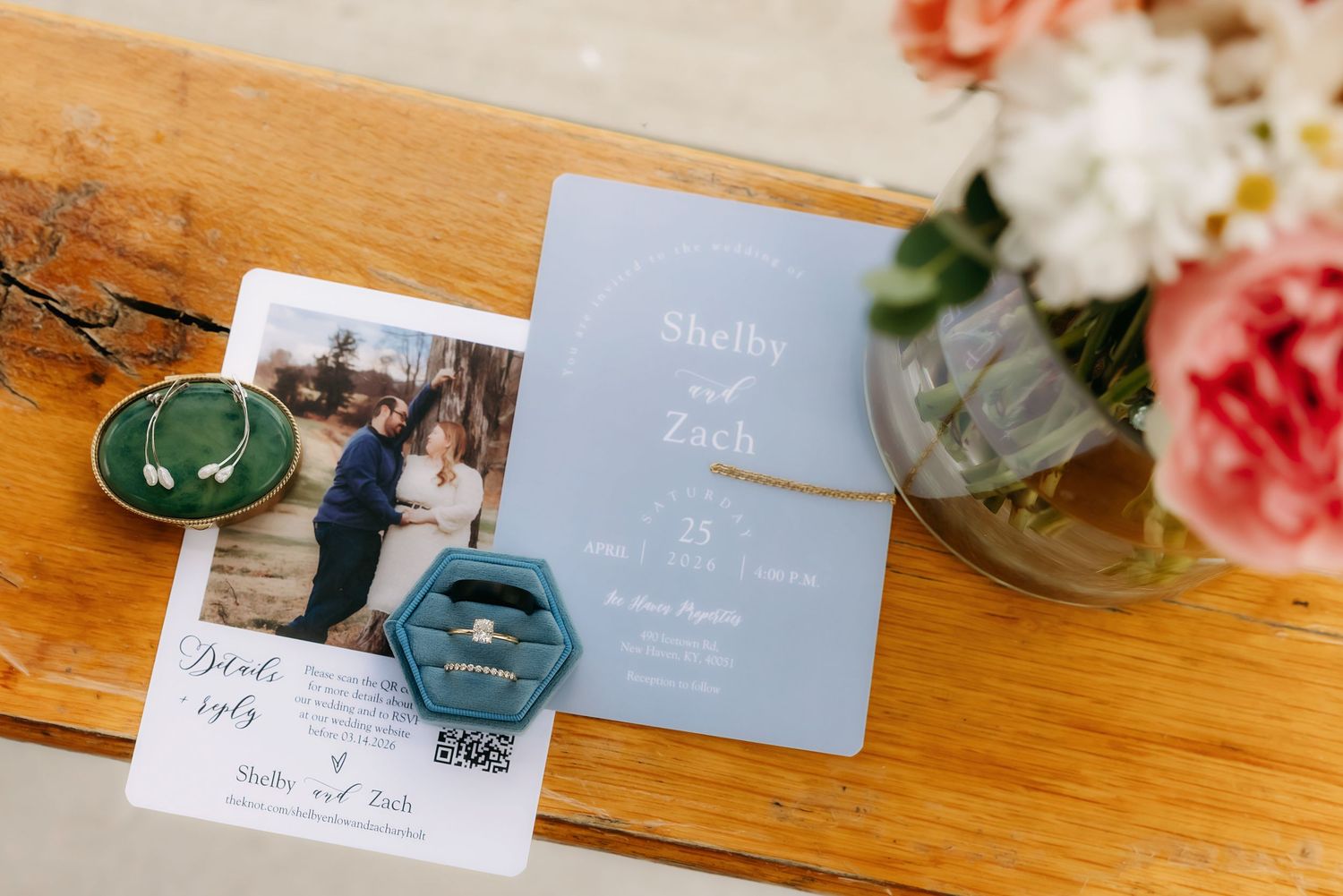 Wedding flat lay with invitation, engagement photo, teal ring box, diamond ring, and floral arrangement.