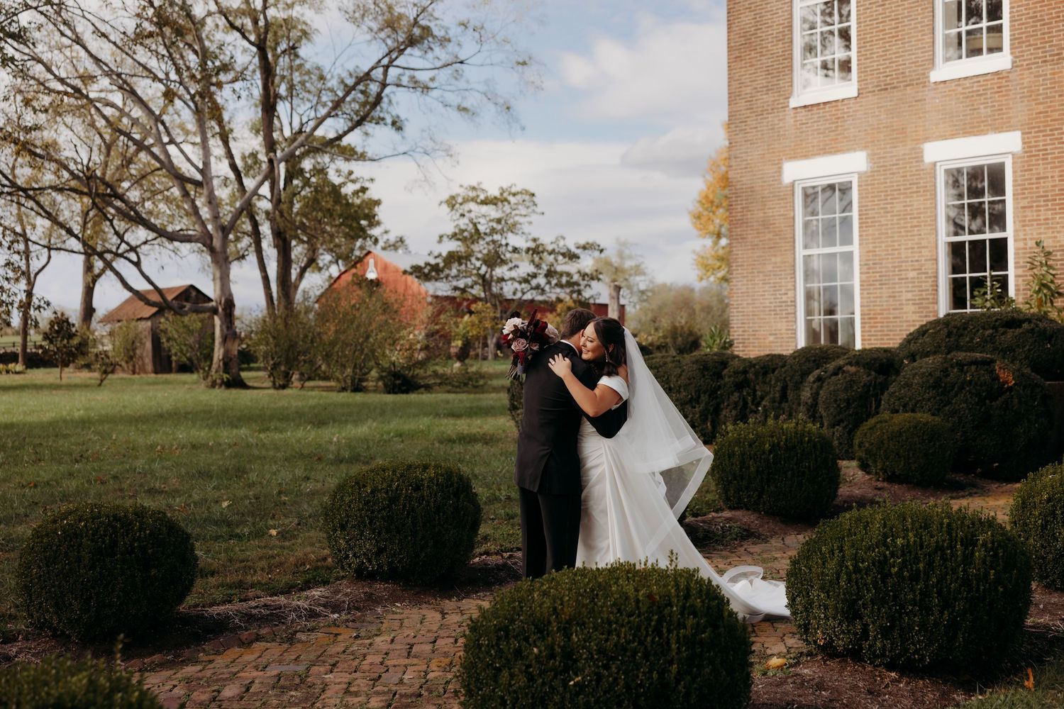 Bride and groom share a romantic kiss in a lush garden beside an elegant brick mansion on their wedding day.