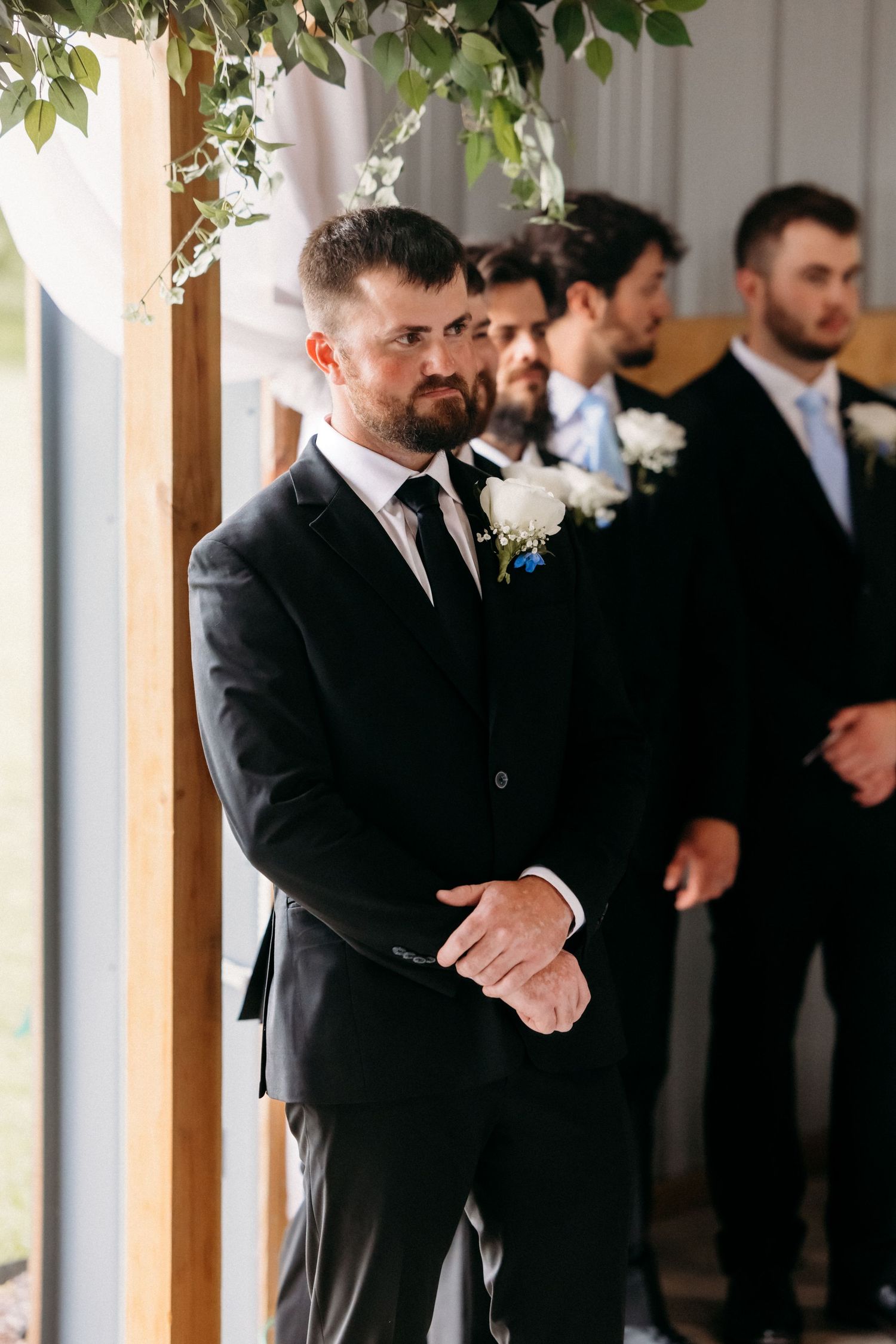 Groom in black suit watches bride walk down aisle flanked by groomsmen at barn wedding.