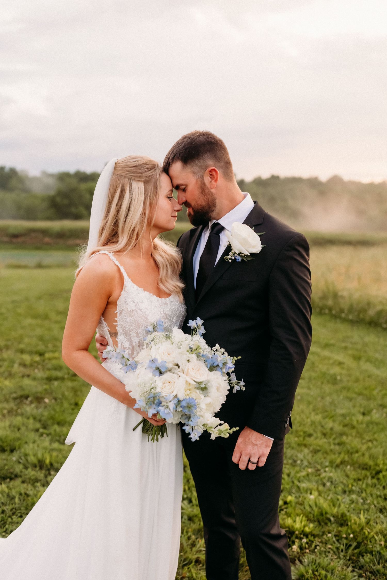 Bride and groom share a romantic forehead touch outdoors, she holds a white and blue bouquet, wearing wedding attire.