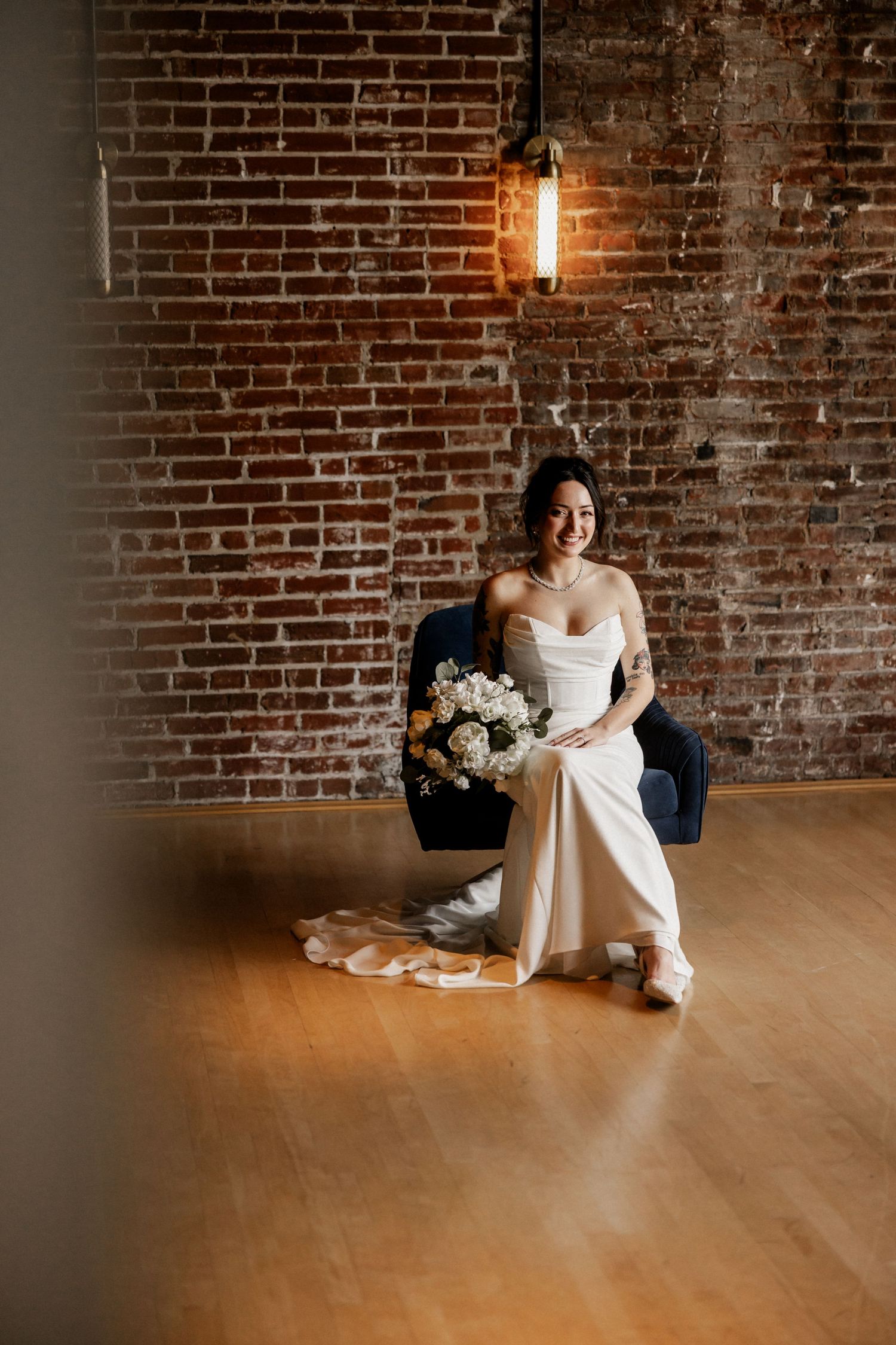 Bride resting on hardwood floor against exposed brick wall with white floral bouquet.