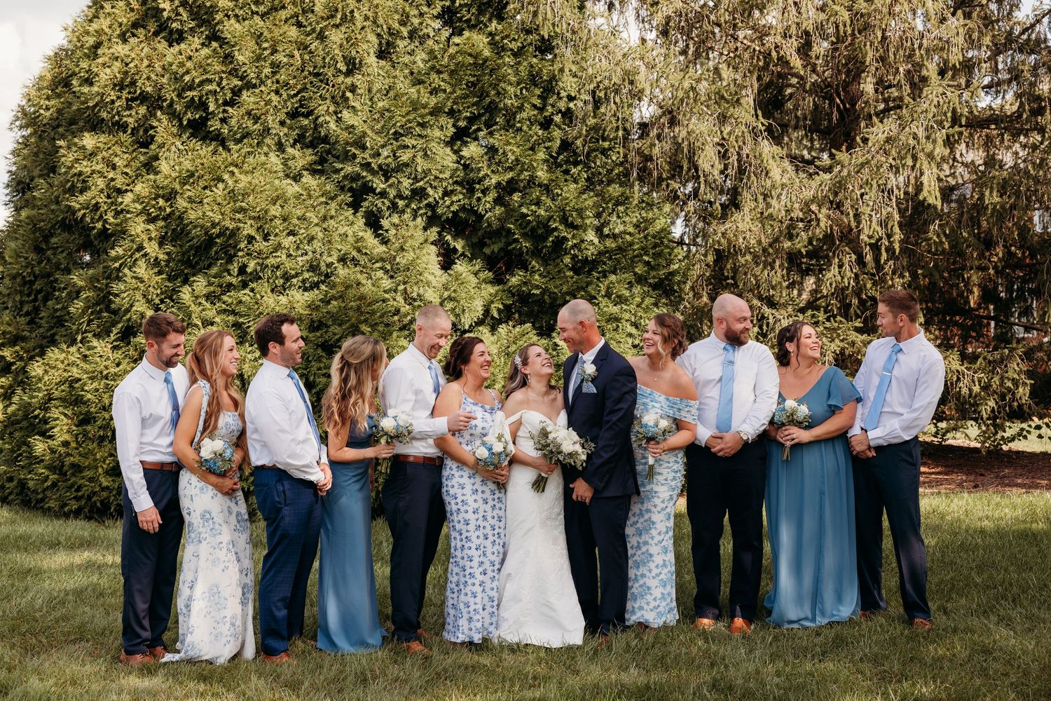 Wedding party group photo on a sunny lawn with trees in the background.