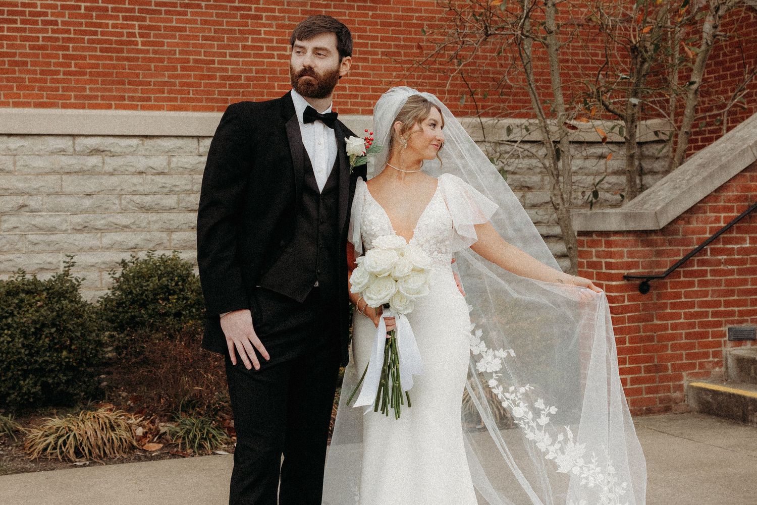 Bride and groom posing outdoors in front of brick wall, bride in white gown with floral veil, groom in black tuxedo.