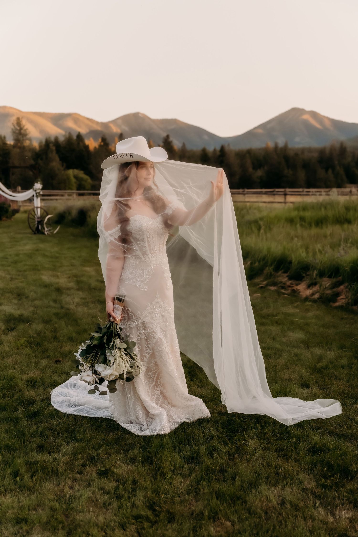 Bride in lace gown, cowboy hat, and flowing veil holds bouquet at sunset with mountain backdrop.
