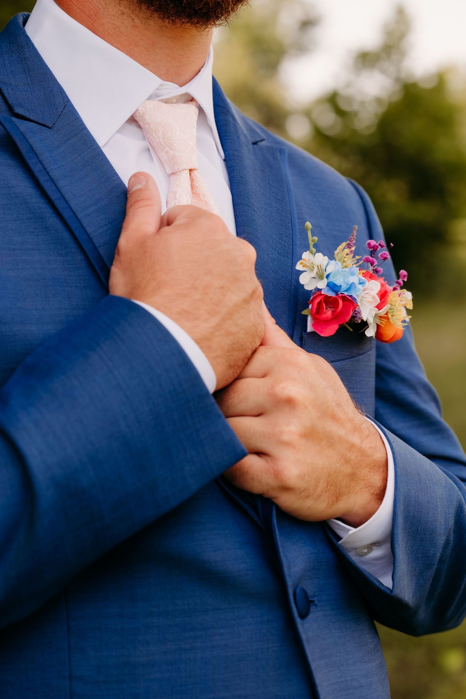 Groom in blue suit adjusting pink tie with colorful floral boutonniere.
