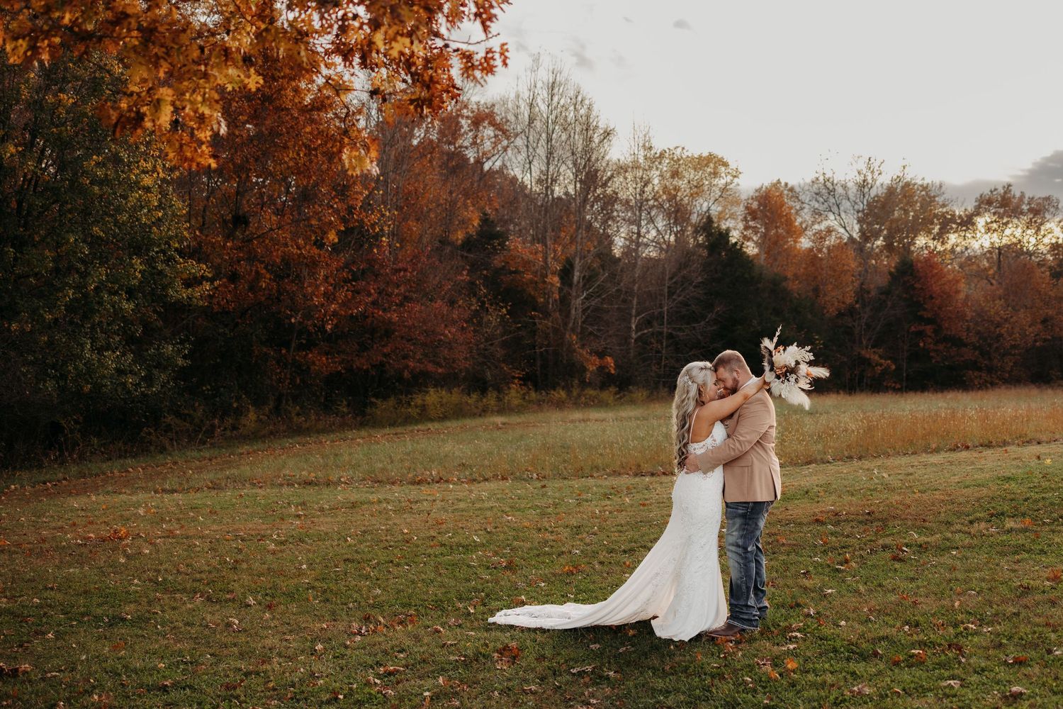 Couple embracing in autumn field with vibrant fall foliage in warm orange and red tones during outdoor wedding.